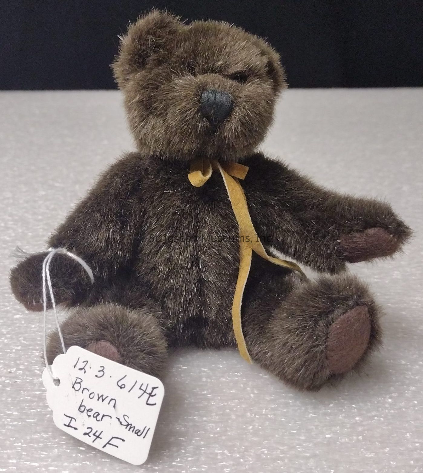 The image depicts a small, plush brown teddy bear with soft fur and a dark brown nose, sitting upright on a textured surface. The bear wears a simple yellow ribbon around its neck, adding a touch of charm to its appearance. Its limbs are positioned in a relaxed manner, with the forearms slightly extended. Attached to the bear's left arm is a white tag, secured with a piece of string, which has handwritten text that reads: "12.3.61 1/4E Brown bear smell I.24 F." This tag suggests that the bear might be part of a collection or inventory system. The background is plain, emphasizing the bear as the focal point of the image, while the lighting highlights the texture of its fur, giving it a cozy and inviting look. The overall composition of the photograph conveys a sense of warmth and nostalgia, reminiscent of cherished childhood memories.