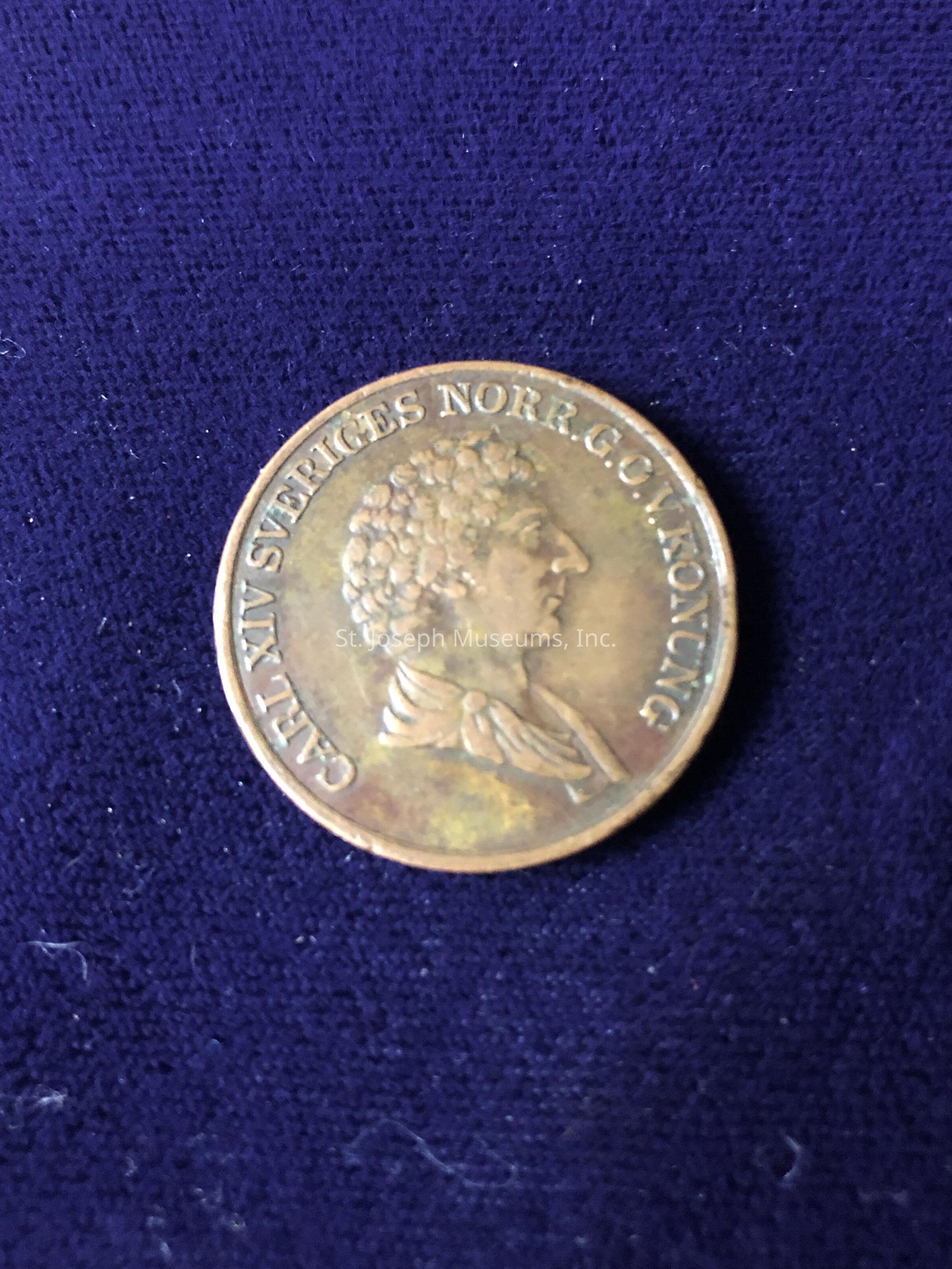 The coin is bronze-toned and displays a profile of a person with curly hair facing to the right. Surrounding the portrait, there is an inscription: "CARL XIV SVERIGES NORR G. O. V. KONUNG." The fabric's rich color contrasts with the warm tone of the metal, highlighting the coin's intricate details and giving it an antique appearance. The overall composition suggests a historical or collectible item, possibly a commemorative or currency piece, reflecting the period's artistry and significance.