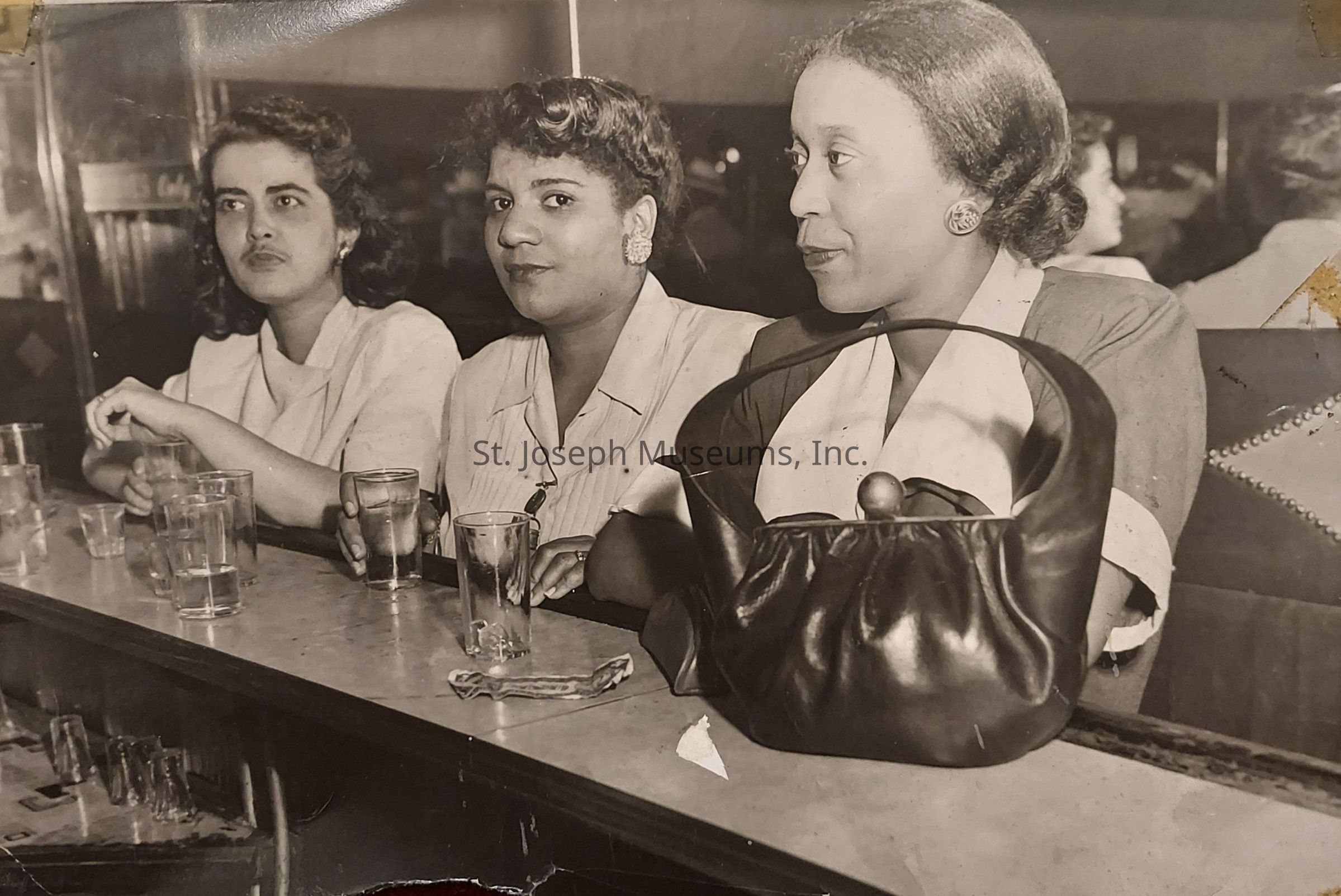 In this black and white photograph, three women are sitting at a bar, each with a glass in front of them, their expressions varying from engaged to contemplative. The woman on the left has dark curly hair and wears a light-colored blouse, while the woman in the middle, with short curls and earrings, seems to be looking directly at the camera. The woman on the right, with her hair neatly styled back, appears thoughtful. In front of her is a large black handbag placed prominently on the bar top.