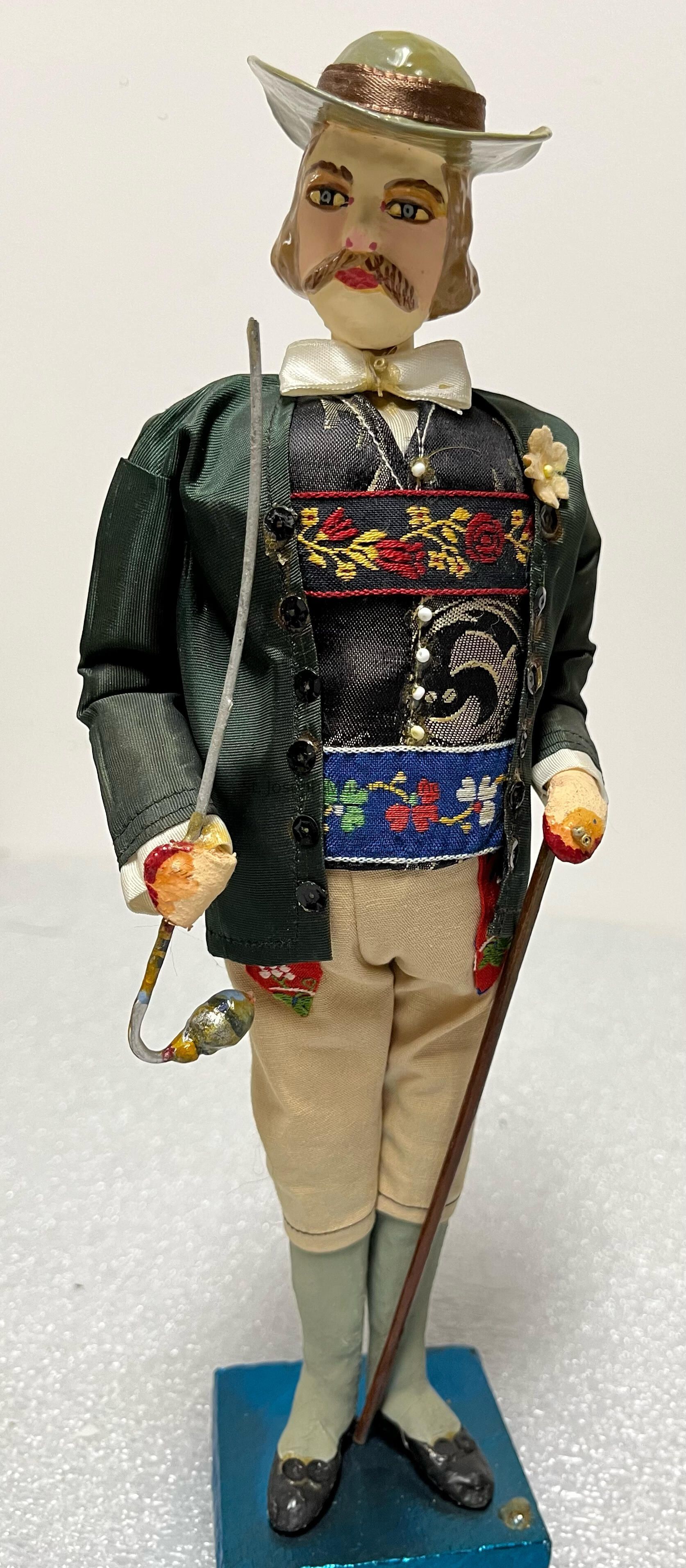 The image depicts a well-crafted figurine of a man dressed in traditional attire, featuring a detailed face with a mustache and striking eyes. He wears a light-colored hat and a bow tie. His clothing includes a dark green jacket with buttons, decorated with intricate floral embroidery in vibrant colors, and matching trousers. He holds a slender sword and wooden staff, adding a sense of historical charm. The figure stands upright on a simple blue base, showcasing a blend of artistry and nostalgia that captures the essence of a bygone era with its attention to detail and cultural representation.