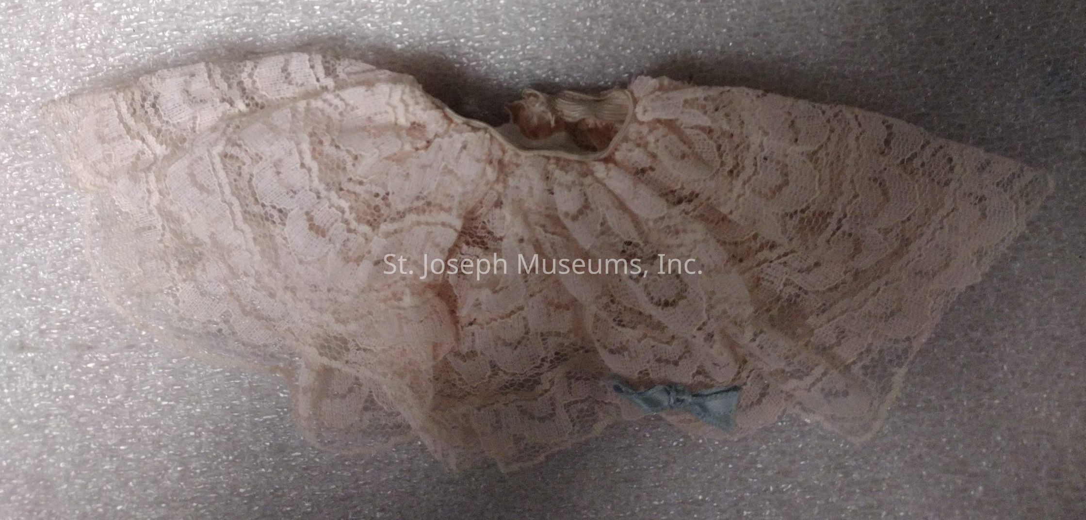 The image displays a delicate, vintage-style baby doll or small dress primarily made from intricate, light pink lace fabric with a floral pattern. The garment has a circular shape, indicating it might be a skirt or part of a dress, with ruffled edges adding to its charm and elegance. Around the collar or waistband area, there seems to be a subtle elastic, allowing for a gathered or bunched look, enhancing its intricate aesthetic. At the bottom corner, a small, light blue ribbon bow adds a touch of contrast and an additional decorative element to the piece. The lace material is sheer, with a texture that appears soft and lightweight, casting gentle shadows on the surface beneath, which resembles a textured, white foam board background, creating a pleasing juxtaposition against the ornate fabric. This playful yet sophisticated design suggests it might be used as clothing for a doll, adding a vintage, timeless elegance to any setting or collection.