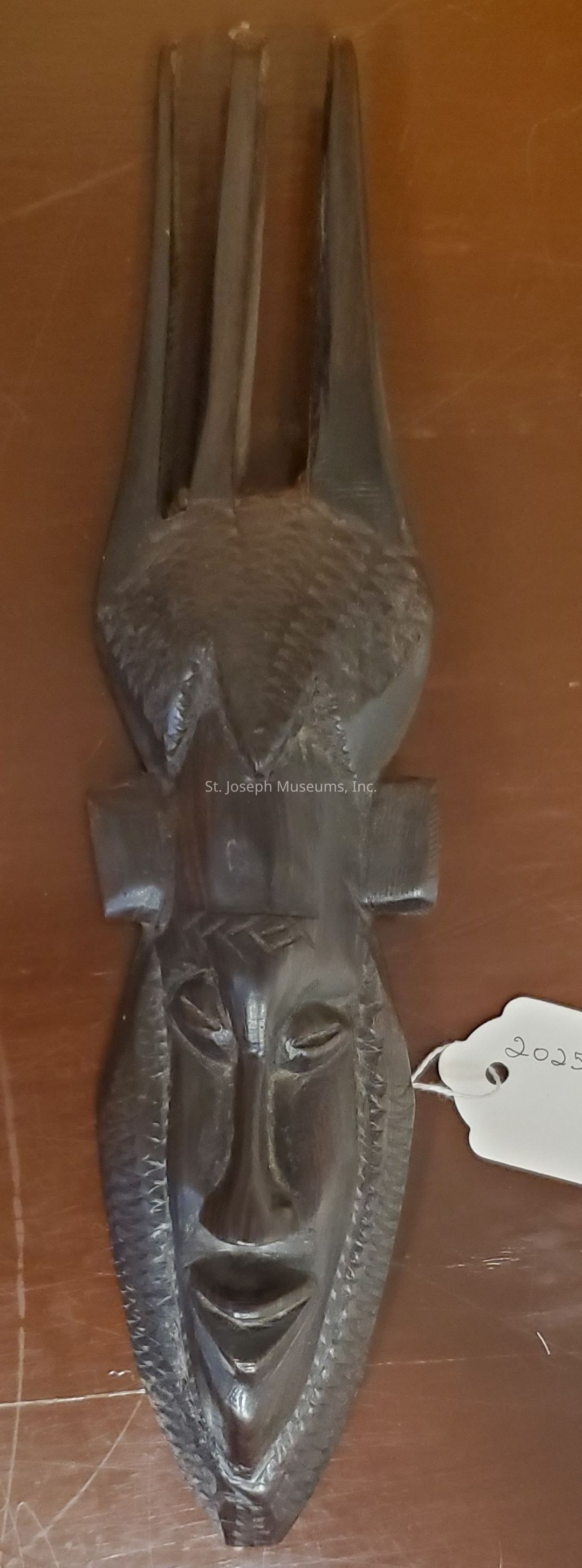 The image depicts a sculpted mask made from dark wood, featuring an elongated face with a serene expression. The mask includes slender eyes, a defined nose, and a slightly open mouth, all intricately carved to provide depth and detail. Above the forehead, two tall, elongated extensions resemble horns or an elaborate headdress, emphasizing the mask’s tall structure. The cheeks contain textured patterns that mimic traditional design elements, enhancing the mask's cultural aesthetic. To the side, a small white tag with the number "2025" is attached, likely indicating an inventory or catalog number. The mask rests against a smooth, brown wooden surface, highlighting its intricate craftsmanship and dark tone in contrast to the lighter background.