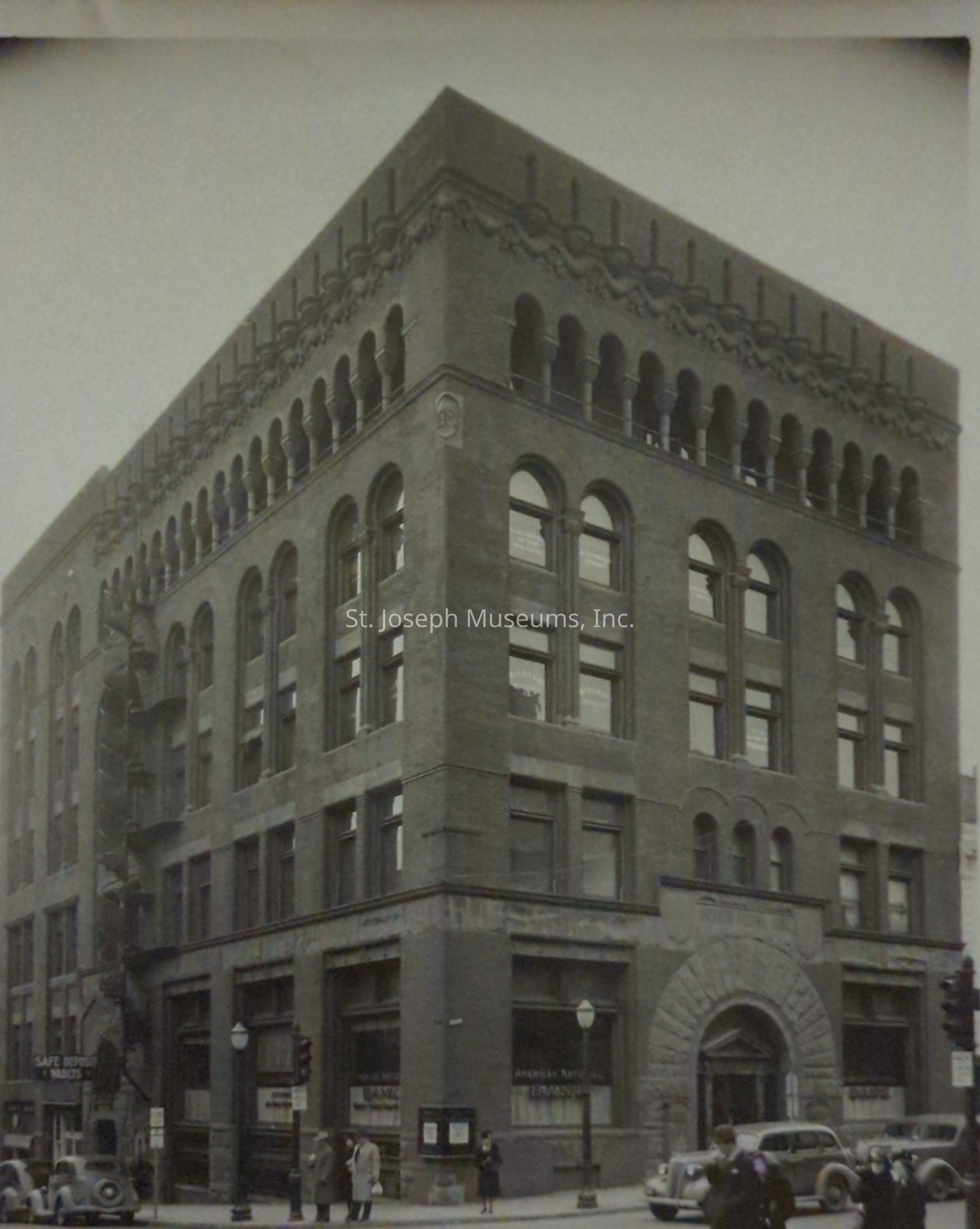 Black-and-white image depicting a historic four-story building on the corner of Felix Street in St. Joseph, Missouri, showcasing early 20th-century architectural design with arched windows, a decorative cornice, and intricate stonework around the entrance. Several cars are parked along the street, and pedestrians, dressed in mid-century attire, are walking along the sidewalks.