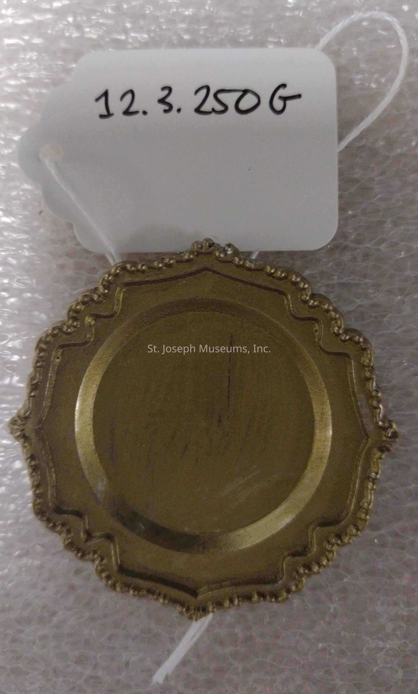 The image shows a decorative, round metal object with an ornate, scalloped edge, possibly a lid or a coaster, placed on bubble wrap. It has a tarnished, aged appearance and is possibly made of brass or similar metal. Attached to it is a white tag with scalloped edges, secured with a thin white string. The tag displays the handwritten numbers and letters "12.3.250G," which could possibly indicate a date and weight or some sort of identification code. The contrast between the textured, protective bubble wrap and the aged elegance of the metal object creates an interesting visual juxtaposition, emphasizing both the item's historical or aesthetic value and the care taken for its preservation.