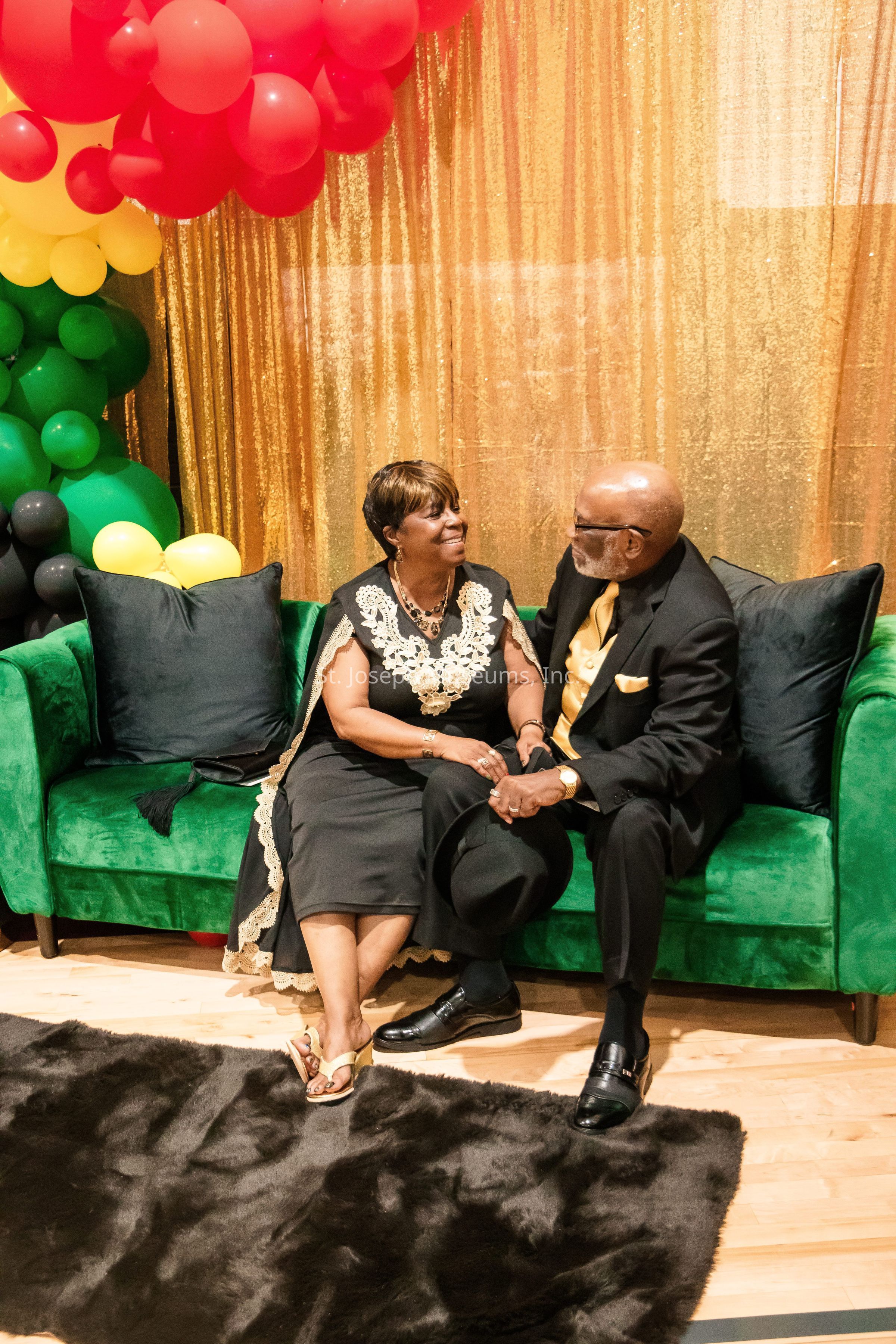 A couple is sitting on a plush green couch, gazing warmly at each other, as the man gently holds the woman’s hand while seated beside her. They are both elegantly dressed; she wears a black dress with lace details and sandals, complemented by a necklace, while he is in a black suit and yellow tie, holding a black hat. The setting is vibrant, with a backdrop of shimmering gold fabric and an arch of colorful balloons in red, yellow, green, and black hues cascading at the side. The couple sits on a wooden floor, and a dark plush rug lies beneath their feet, creating a cozy and inviting atmosphere. This heartwarming scene suggests a moment of celebration or a formal gathering, radiating joy, affection, and a strong connection between the two individuals. The scene is beautifully composed, balancing elegance and festivity, with the soft lighting adding warmth to the already intimate setting.