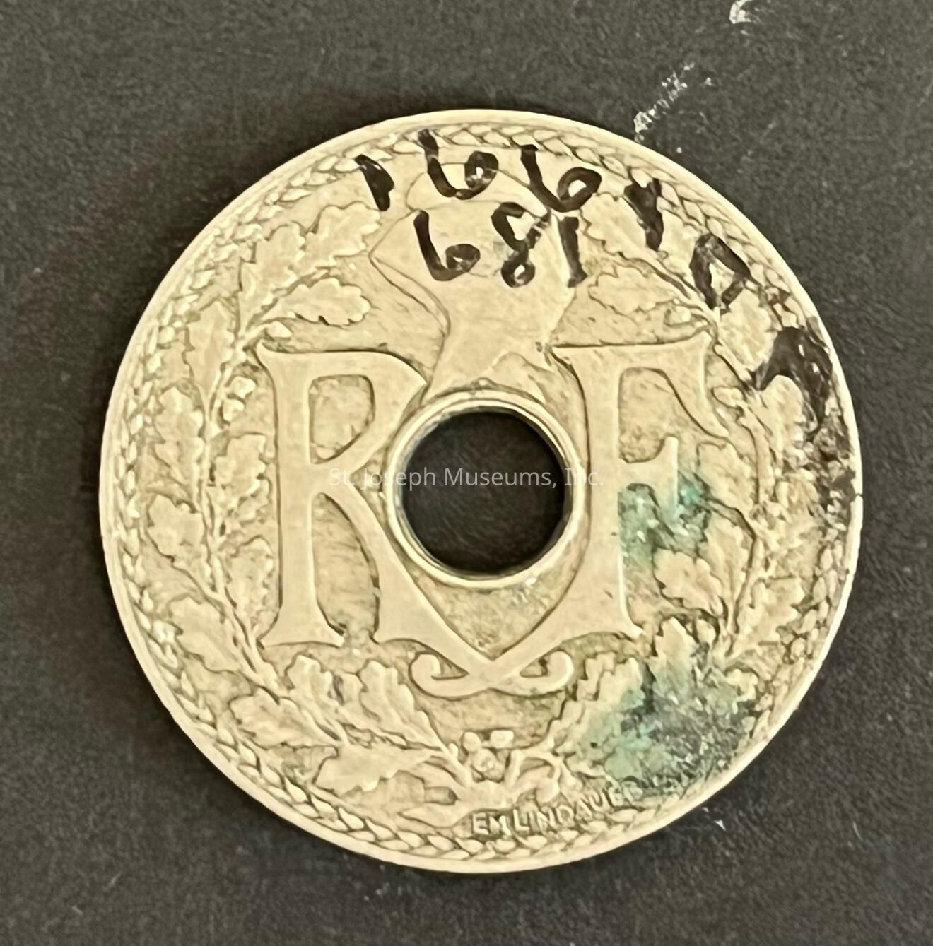 The image features a vintage coin with a circular hole in the center. The coin is adorned with intricate, leafy patterns along its edge, creating a decorative border. Prominently displayed in the center are the large initials "RF," which stand out against the textured background. The coin has visible aging marks, including some discoloration and tarnish, particularly near the bottom right area. Above the initials, handwritten numbers "1916" and "1891" are visible, possibly indicating dates. The text "EM LINDAUER" appears at the lower part, likely denoting the designer or mint. The coin's design suggests it might be a historical French piece, given the "RF" traditionally represents "République Française." The overall appearance of the coin, with its elegant motifs and signs of wear, reflects its historical and numismatic significance. The coin is laid against a dark surface, emphasizing the details and craftsmanship of the design while highlighting areas of wear and aging in the metal.