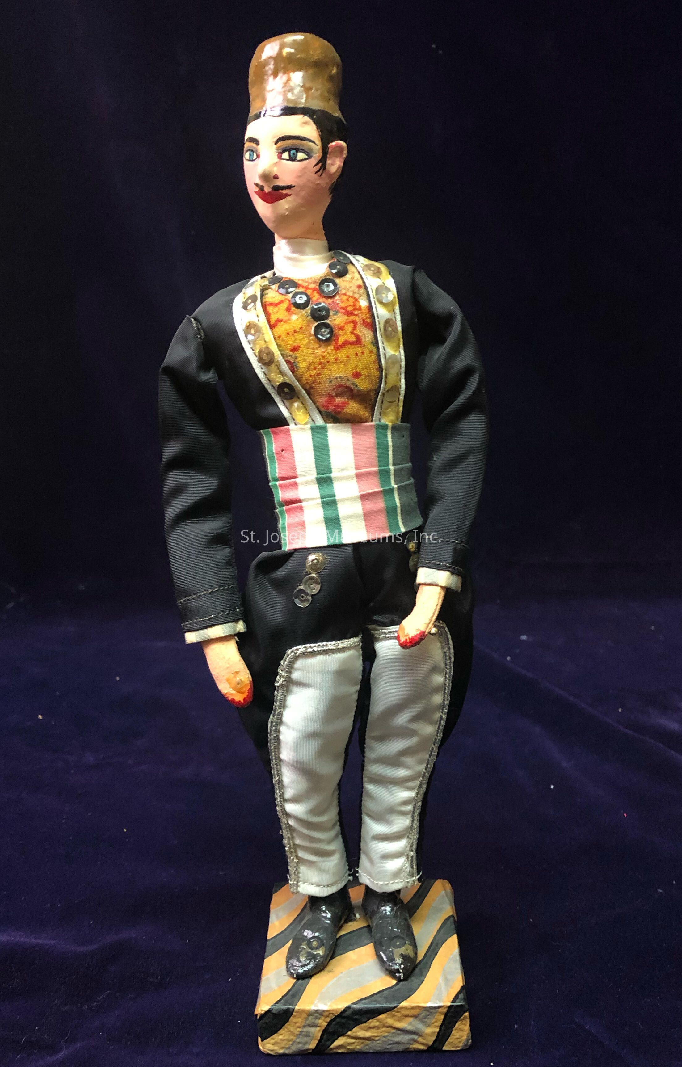 The image portrays a traditional doll standing upright against a dark background, showcasing detailed craftsmanship, with the doll dressed in a richly ornamented costume featuring a black jacket adorned with decorative buttons and elaborate embroidery. The outfit also includes white trousers with silver detailing, and a colorful, striped sash around the waist. The doll's face is painted with vivid features including bright eyes and a mustache, topped by a dark hat. It stands on a square base painted with a striped pattern, giving it a sturdy appearance. The intricate details suggest cultural significance, likely representing traditional or historical attire.