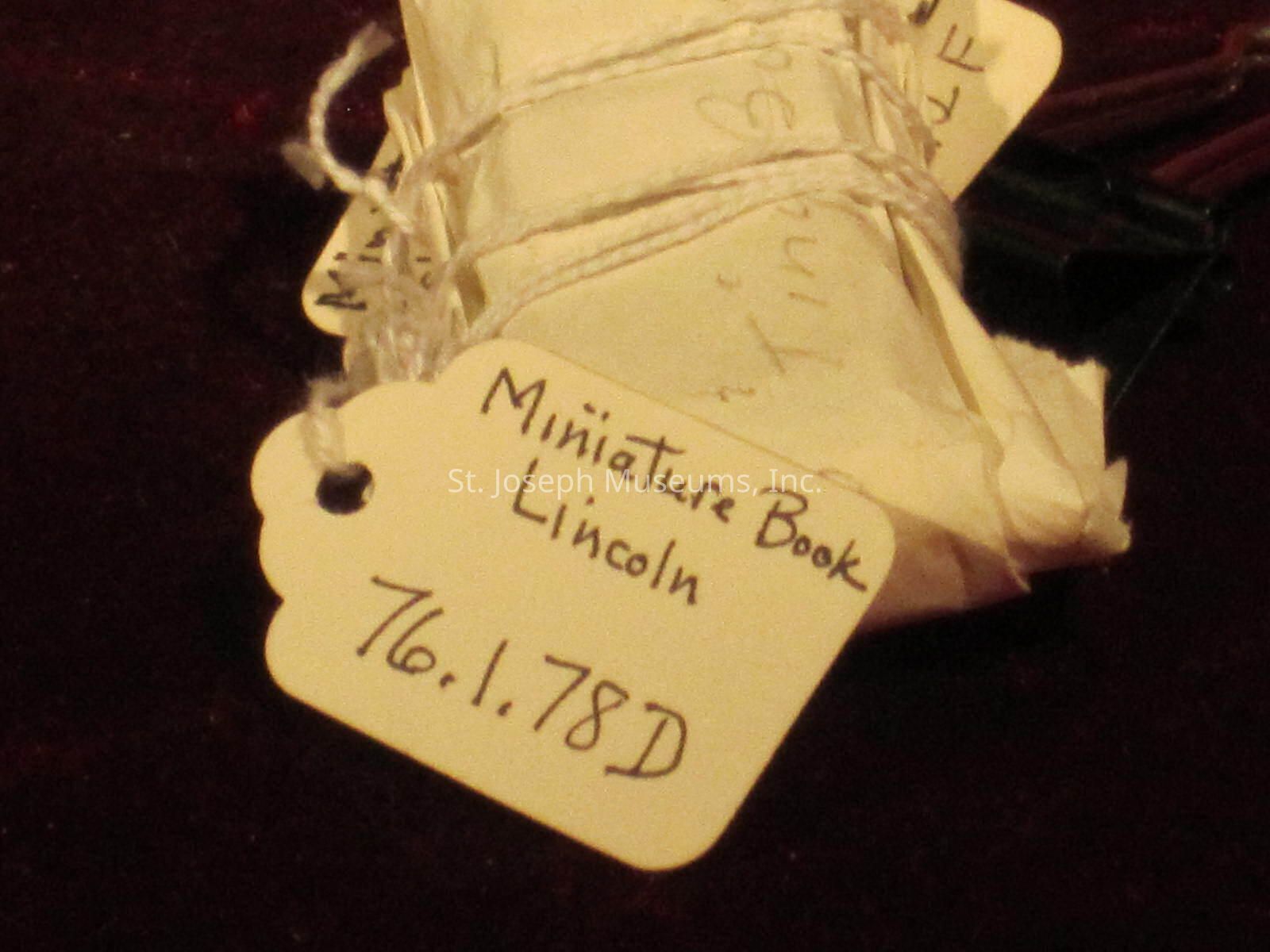 The image shows a small, wrapped object with a scalloped-edged tag attached. The tag is labeled "Miniature Book Lincoln" and "76.1.78D," indicating some form of cataloging or identification number. The wrapping is tied with white string, suggesting a careful and possibly archival method of preservation. The background is dark red, which contrasts with the white wrapping and tag, enhancing the focus on the object. The handwritten text on the tag gives it a personal or historical feel, hinting that it might be part of a collection or exhibit potentially related to Lincoln-themed artifacts or memorabilia.
