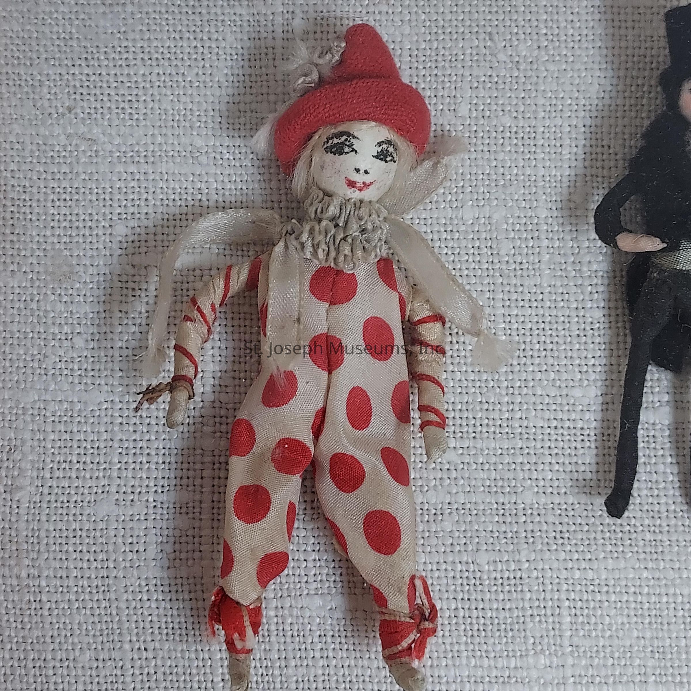 The image depicts a small clown doll lying on a textured white surface, featuring a whimsical, handcrafted appearance. The doll is adorned with an off-white costume prominently displaying large red polka dots that catch the eye. Complementing the cheerful attire, the clown wears a matching red hat with a jaunty tip, adding to its playful charm. Its face is intricately painted, showcasing exaggerated makeup typical of clowns, with dark eyes, raised eyebrows, and a wide red smile. The neck of the doll is decorated with a ruffled collar made from a translucent material, enhancing its vintage aesthetic. The limbs are simple and slender, with arms slightly raised, suggesting a playful pose. To the right, part of another figure is visible, dressed in black with a similar fabric design, creating a sense of a whimsical collection or theme. The craftsmanship exhibits delicate attention to detail, evoking nostalgia and the joyful essence of classic circus clowns.