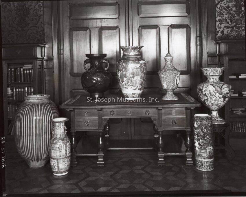 Tootle Mansion Vases, c. 1900: A black and white photograph depicting a collection of intricately designed vases arranged on a sturdy wooden table in the Tootle Mansion, located at 11th & Charles Street in St. Joseph, Missouri. These vases, each distinct in shape and decoration, are made from various materials, showcasing both intricate patterns and textures. The background features an elegant wooden paneling that enhances the historical and artistic ambiance of the interior.