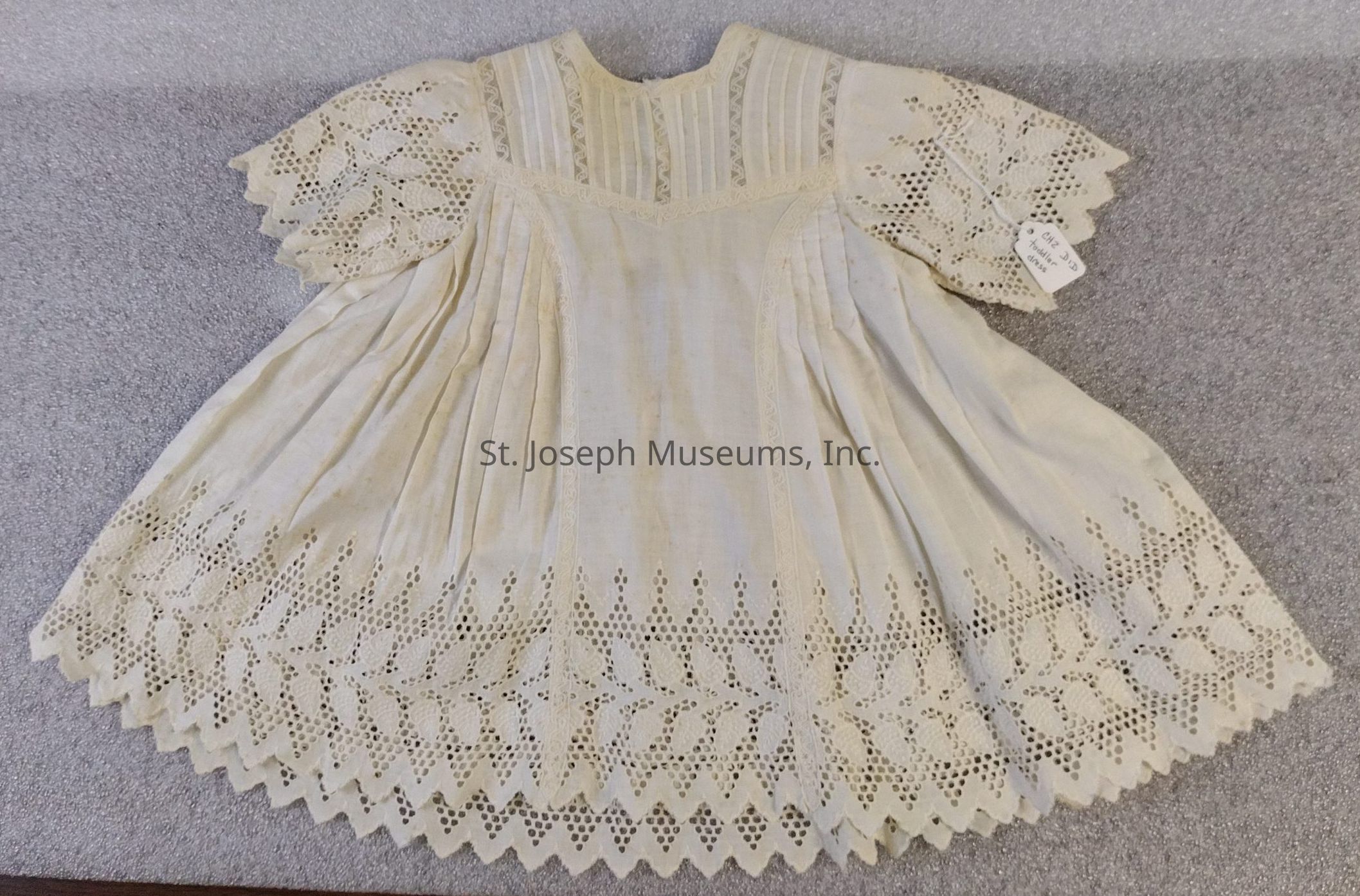 The image shows a toddler-sized white cotton dress, designed to be knee-length, featuring eyelet lace detailing at the hem and sleeves. Lace strips decorate the yoke and extend down the front of the dress. The dress is adorned with an array of pleats and a scalloped edge at the hem. The lace on the sleeves forms a floral pattern.