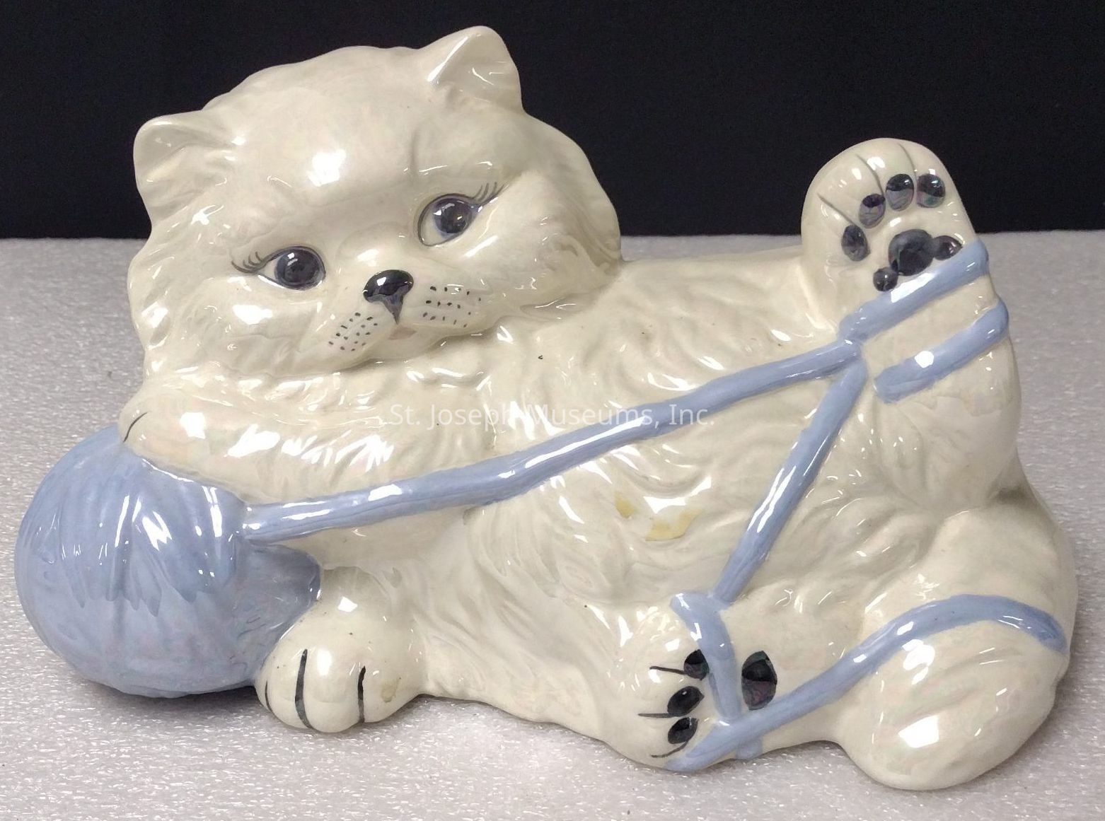 The image depicts a ceramic figurine of a playful, fluffy cat lying on its back with a content expression on its face. The cream-colored cat has big, expressive eyes and realistic details on its fur and whiskers, adding a touch of charm. It is holding a skein of yarn, light blue in color, that is playfully wrapped around its paws and legs, suggesting a moment of mischievous entertainment. The cat’s one paw is raised up, with visible painted paw pads, while its body is positioned in a relaxed, lounging pose. The glossy finish of the ceramic reflects light, emphasizing the smoothness of the figure. Set against a neutral background, the cat and yarn stand out, creating an inviting, whimsical scene that evokes a sense of warmth and coziness. This figurine could easily serve as a delightful decorative piece that captures the playful nature and curiosity of a cat, making it a charming addition to any collection.