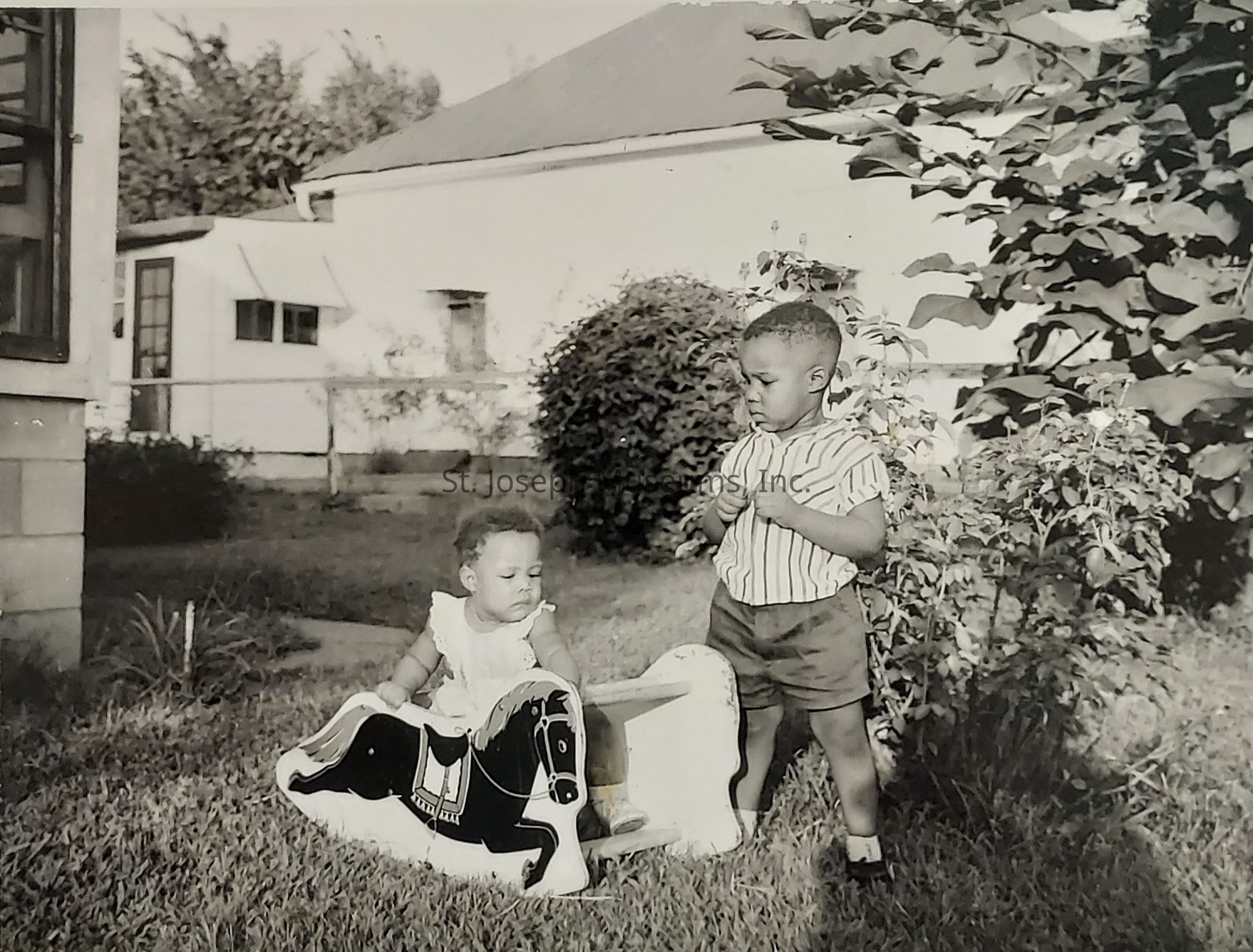 In a vintage black and white photograph, two young children are seen playing in a yard, capturing a moment of childhood innocence and exploration. The scene is set in a grassy area with a house and shrubbery in the background. The older child, standing to the right, is dressed in a striped short-sleeve shirt and dark shorts, and seems engaged in an activity, possibly deep in thought or in conversation. The younger child sits on a playful, decorative piece shaped like a horse, focused intently on it, dressed in a light-colored outfit. The image exudes a sense of nostalgia, emphasizing the simple joys and curiosities of childhood in a suburban setting, framed by the rich textures of the foliage and architecture around them, indicative of a family home environment.