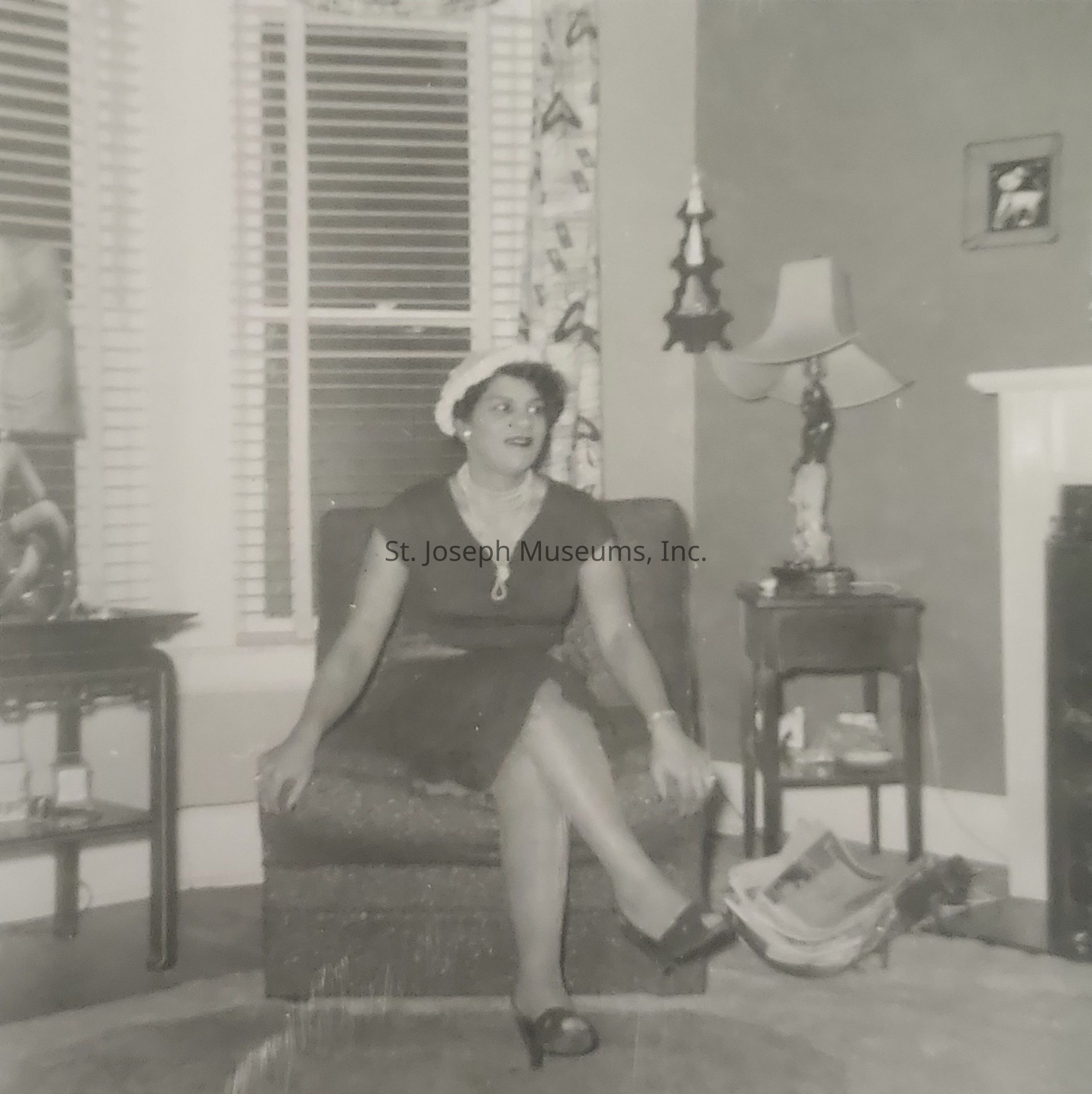 The image depicts a vintage setting inside a living room, where a woman is seated gracefully on an armchair, her posture relaxed but poised as she looks slightly off to the side, wearing a dark dress complemented with a light-colored hat and necklace, adding a touch of elegance to her ensemble; her legs are crossed, and she is wearing dark heels. The room is tastefully decorated with patterned curtains drawn over a window with blinds, a small side table nearby holding a decorative lamp with a figurine base, while newspapers are casually placed on the floor beside it, suggesting a cozy and lived-in environment. A framed photo is hung on the wall above a mantelpiece, indicating a personal and warm atmosphere typical of mid-20th-century homes.