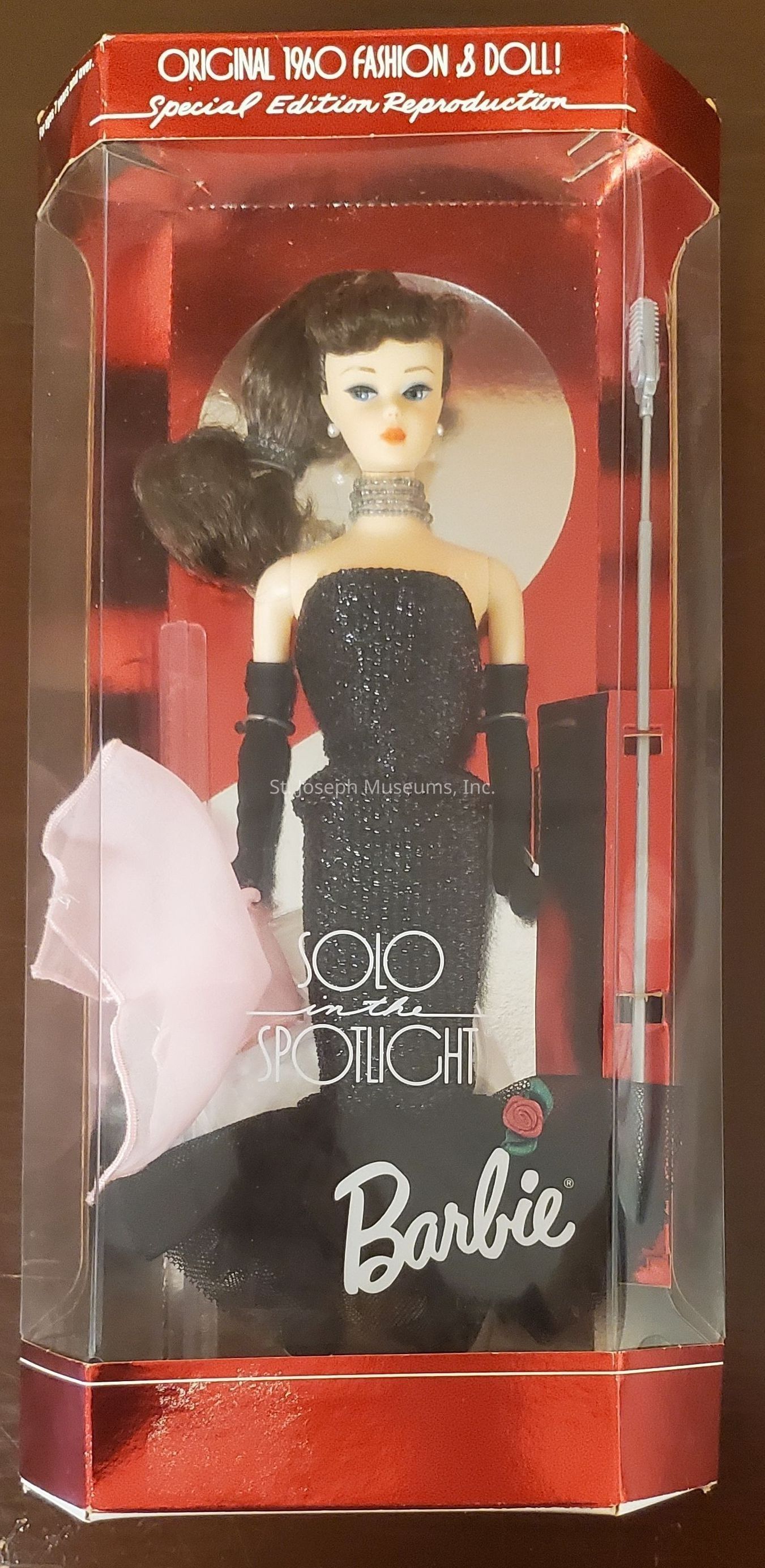 The image shows a "Solo in the Spotlight" Barbie doll encased in its original packaging, which is a special edition reproduction reflecting the original 1960 fashion and doll design. The packaging is primarily red with clear plastic allowing a full view of the doll inside, which is elegantly dressed in a shimmering black gown, accessorized with long black gloves and pearl earrings. The doll's packaging also includes a microphone accessory on the right and a pink scarf on the left, emphasizing a glamorous performance theme. The front of the box prominently displays the Barbie logo along with a red rose design, enhancing the classic and collectible appeal.