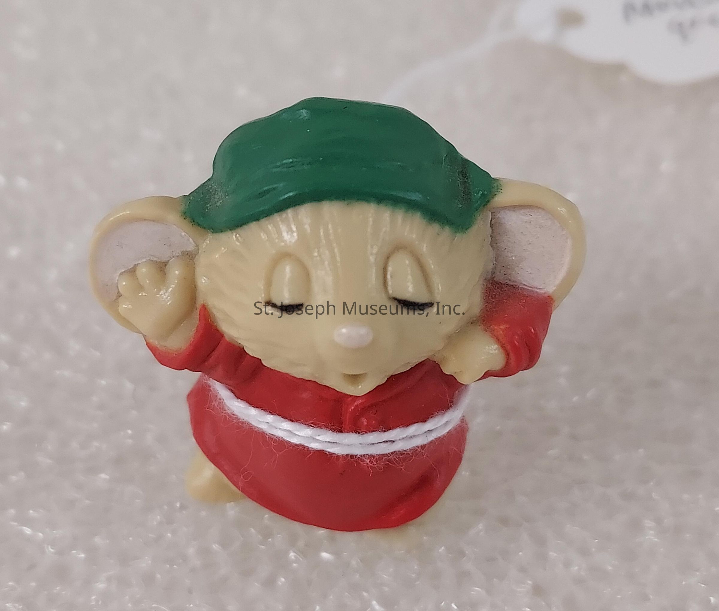 The image features a small, adorable figurine of a mouse, wearing a bright red robe tied with a white sash around its waist and a dark green cap on its head, set against a textured background. The mouse is depicted with its eyes closed, giving the impression it is either in a state of relaxation or deep thought. Its large, rounded ears are prominently displayed, and one of its small arms is raised, creating a charming and playful demeanor. The simplicity of its design, coupled with the soft colors, lends the figurine a cute and whimsical appearance. The mouse figurine appears to be standing on a surface that has a grainy, snowy texture, adding an element of warmth and coziness to the overall scene. The attention to detail is evident in the folds of the robe and the serene expression on the mouse's face, suggesting careful craftsmanship. The figurine could be part of a collection or a decorative item meant to evoke a sense of comfort and joy.