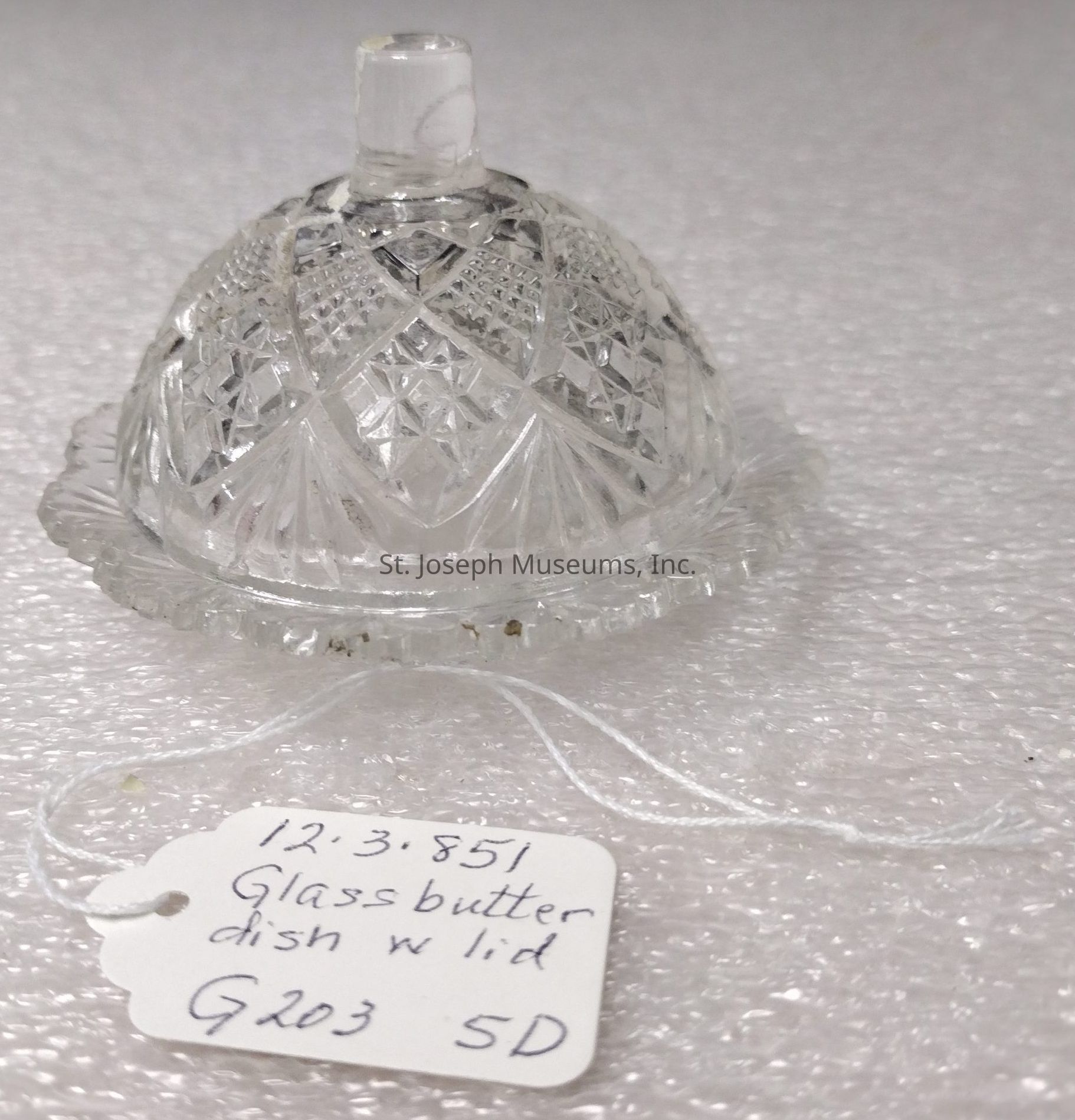 The image shows a clear glass butter dish with a lid, featuring intricate cut glass patterns that create a decorative and elegant appearance. The lid is dome-shaped and has a small handle on top for easy lifting. The dish rests on a round base with a slightly ridged edge, adding to its ornate design. Attached to the dish is a white tag with handwritten text identifying it as a glass butter dish with lid, and providing some notation, possibly an inventory number, which reads "12.3.851" and additional identifiers "G203 5D." The background appears to be a textured white surface, possibly a protective layer for display or storage, which contrasts with the clear, reflective quality of the glass, enhancing the overall aesthetic of the piece.