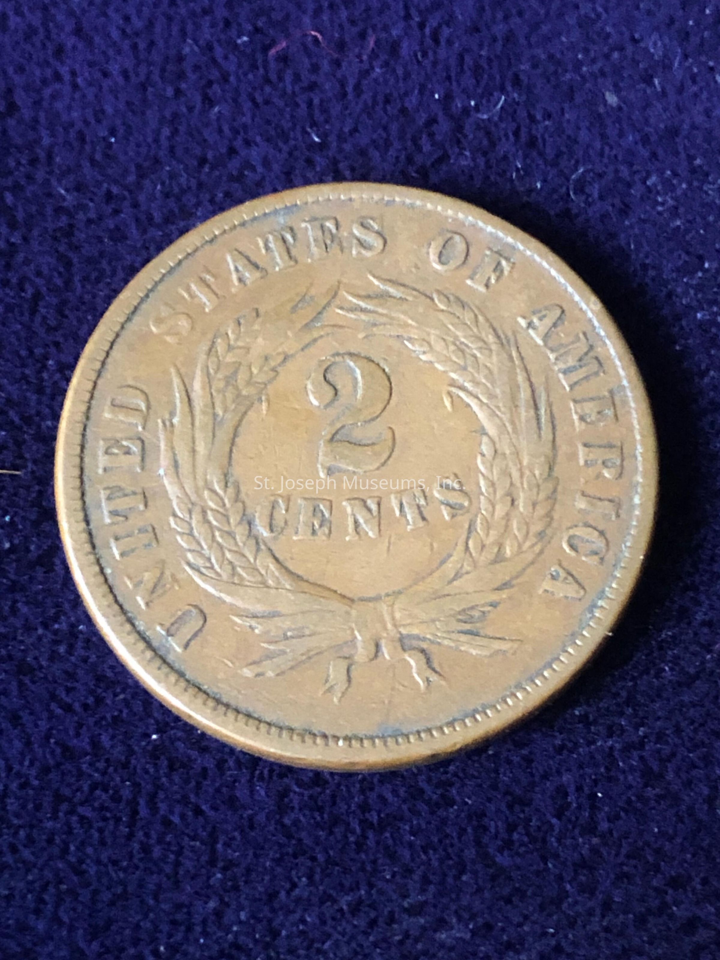The image shows an antique coin from the United States of America, specifically a two-cent piece, which is prominently marked with the denomination "2 CENTS" in the center, surrounded by a wreath. The text "UNITED STATES OF AMERICA" encircles the outer edge of the coin, which features a classic, worn patina indicative of its age, against a dark purple velvet background that enhances its historical and numismatic significance.