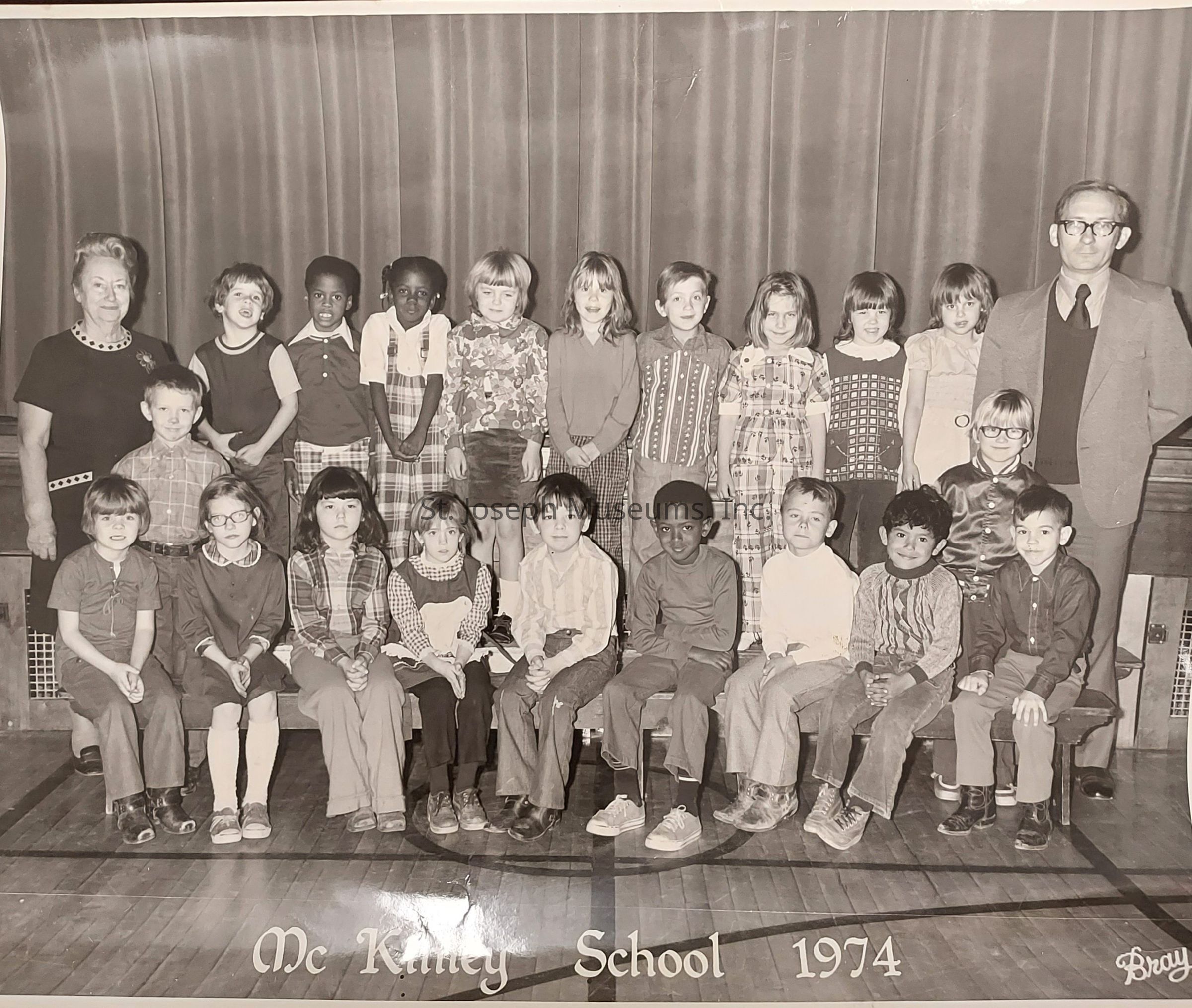 The image depicts a black-and-white class photo from 1974 at McKinley School, showcasing a group of young children along with two adults, likely their teachers, posed in front of a curtain backdrop. The children, positioned in two rows, display a variety of attire typical of the 1970s, including plaid patterns, dresses, and vests, with some students sitting and others standing. The adults, standing on either end of the group, are dressed in more formal attire, with one woman wearing a dress and a man in a suit and tie. The scene emanates a nostalgic and historical vibe, capturing a moment in time within an educational setting.