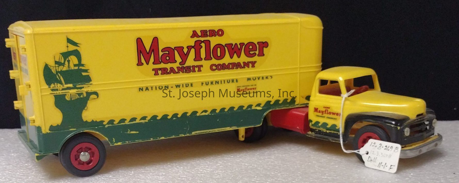 The image shows a vintage toy truck, modeled after an old moving truck, painted in vibrant yellow with red accents. The vehicle is a representation of the "Aero Mayflower Transit Company," as indicated by the bold red text on the side of the yellow cargo section. A dark green graphic of a ship under full sail is silhouetted on the side, complementing the company's theme. The toy features a cab, also predominantly yellow, with a contrasting black bumper and wheel arches, and the wheels are red with silver centers. A small, white tag is attached to the front of the truck, inscribed with numbers and text.