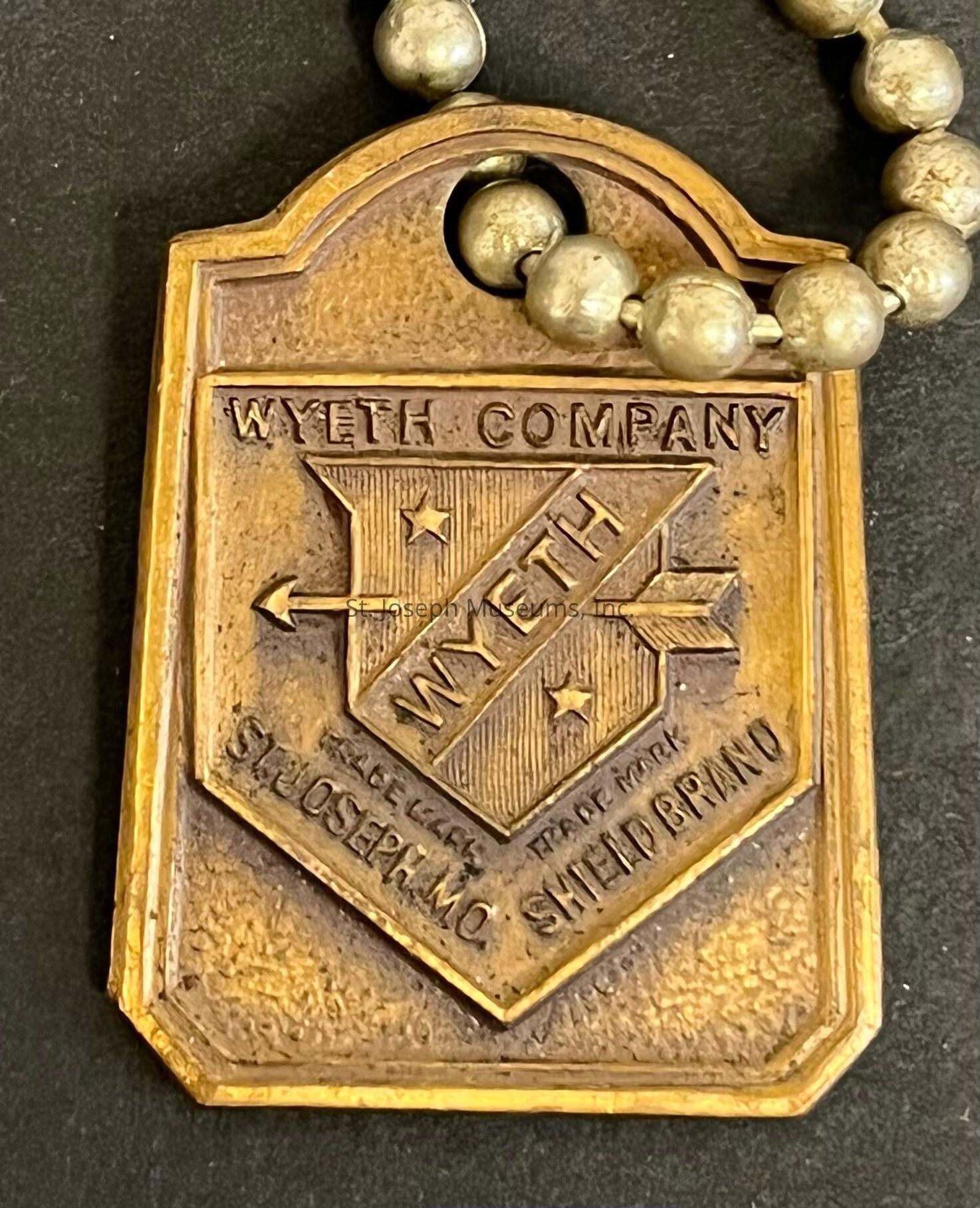 The image displays a vintage brass tag with a rectangular shape and rounded top edges, featuring an embossed design that reads "WYETH COMPANY" and "MYETH" in a stylized font across a banner overlaid on a shield emblem with two arrows and stars, accompanied by the engraved words "ST. JOSEPH, MO." and "SHIELD BRAND," indicative of a historic branding or trademark element, all attached to a string of metal beads, suggesting it may be part of a piece of industrial or promotional memorabilia, possibly used for identification or marketing purposes in a bygone era, with the patina and wear on the surface suggesting age and historical significance, further enhanced by the intricate detailing and robust style commonly seen in early to mid-20th-century industrial artifacts, hinting at its origin from a period where such tags played a pivotal role in branding and identity representation, giving it a strong aesthetic and nostalgic appeal to collectors or those with an interest in industrial history.