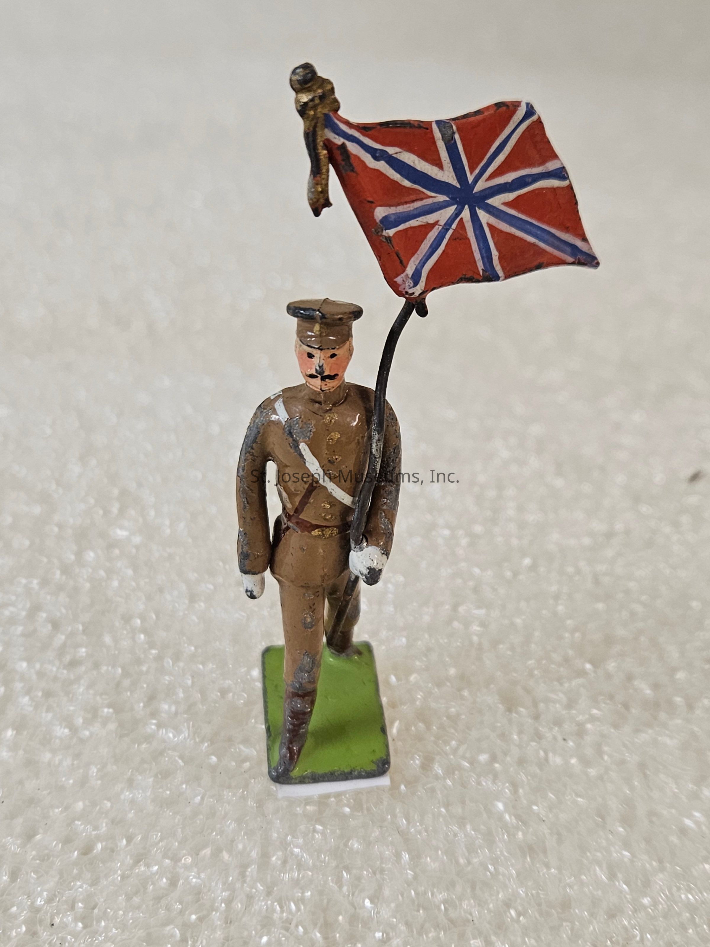 The image depicts a small, vintage figurine of a soldier in a brown uniform, complete with a peaked cap and boots, standing upright on a flat green base. The soldier is holding a Union Jack-inspired flag with a metal pole, which features a red field intersected by white and blue lines resembling the iconic design. The figurine appears to be made of painted cast metal, evident by the slightly worn state of the paint, revealing some wear and tear, giving it a vintage or antique appearance. The background consists of a textured, light-colored surface that impacts the lighting and shadows, creating a neutral, soft effect behind the figurine. The flag and uniform suggest a mid-20th-century style, hinting at a historical or collectible theme, possibly linked to military history or toy soldier collections. The craftsmanship demonstrates attention to detail despite the small scale, reflecting both artistic and historical elements.