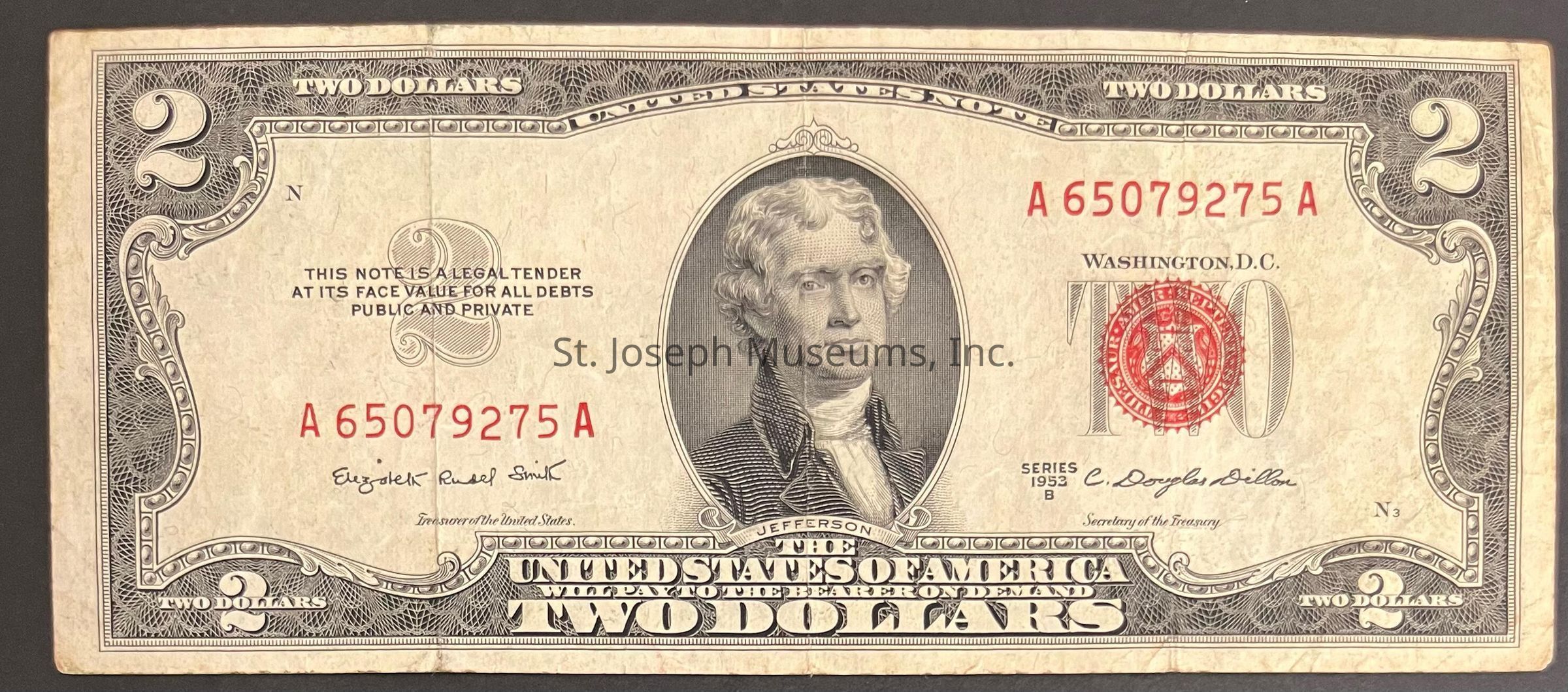 The image depicts a United States two-dollar bill from the series of 1953 B. The bill features an intricate border design and a red seal on the right side. In the center, there is a prominent portrait of Thomas Jefferson, the third President of the United States, who is framed within an oval design. The serial number "A 65079275 A" is printed in red ink above the portrait, with smaller ornate text stating the denomination of the bill at the top and bottom. Below Jefferson's portrait, the name "Jefferson" is printed, and to the left, the words "THIS NOTE IS A LEGAL TENDER AT ITS FACE VALUE FOR ALL DEBTS PUBLIC AND PRIVATE" appear. At the bottom, the text "UNITED STATES OF AMERICA," flanked by "TWO DOLLARS," is prominently displayed. The signatures of the Treasurer and Secretary of the Treasury are visible, alongside the phrase "In God We Trust" and other official inscriptions associated with