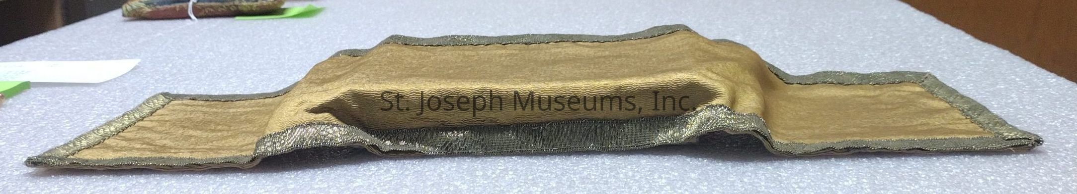The image displays a flat, ornate object with a central raised section, crafted from a shiny, gold-colored fabric that has an intricate texture. This fabric object has metallic trim or border, adding to its decorative appearance. It is laid out on a textured, light gray surface that resembles a table or countertop with a fabric-like covering. In the background, there are blurred elements including a green object with a white string and what appears to be a piece of paper, partially out of focus, contributing to a sense of depth within the image. The ornate object's specific use is not directly evident from the image, but its elaborate and crafted nature suggests it could be a type of decorative textile, perhaps used in a cultural or ceremonial context, or as a decorative covering. Its appearance of being padded or cushioned implies it might be intended to hold or protect something delicate.