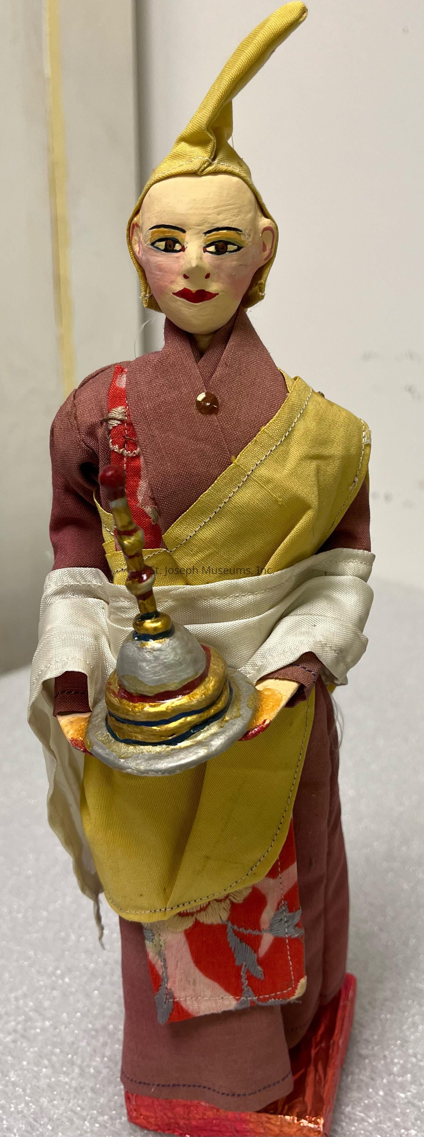 The image shows a figurine dressed in traditional attire, featuring a fabric combination of red, yellow, and white with intricate patterns, and holding an ornamental object. The figure has a pale face with painted features, including black hair styled in an upright manner, yellow eyeshadow, red lipstick, and earrings. The clothing consists of a red robe with a vertical collar, overlaid by a yellow sash draped across the left shoulder and secured at the waist. The arms are wrapped in white fabric, creating an elegant appearance. The base is adorned with red and pink floral patterns. In the figure's hands rests a small circular tray displaying a sculpted representation of what appears to be a traditional ceremonial offering, artistically detailed with gold, red, and black paints. The overall design and attire of the statuette suggest inspiration from cultural or religious themes, capturing intricate craftsmanship and cultural symbolism, evoking an aura of history, tradition, and artistry.