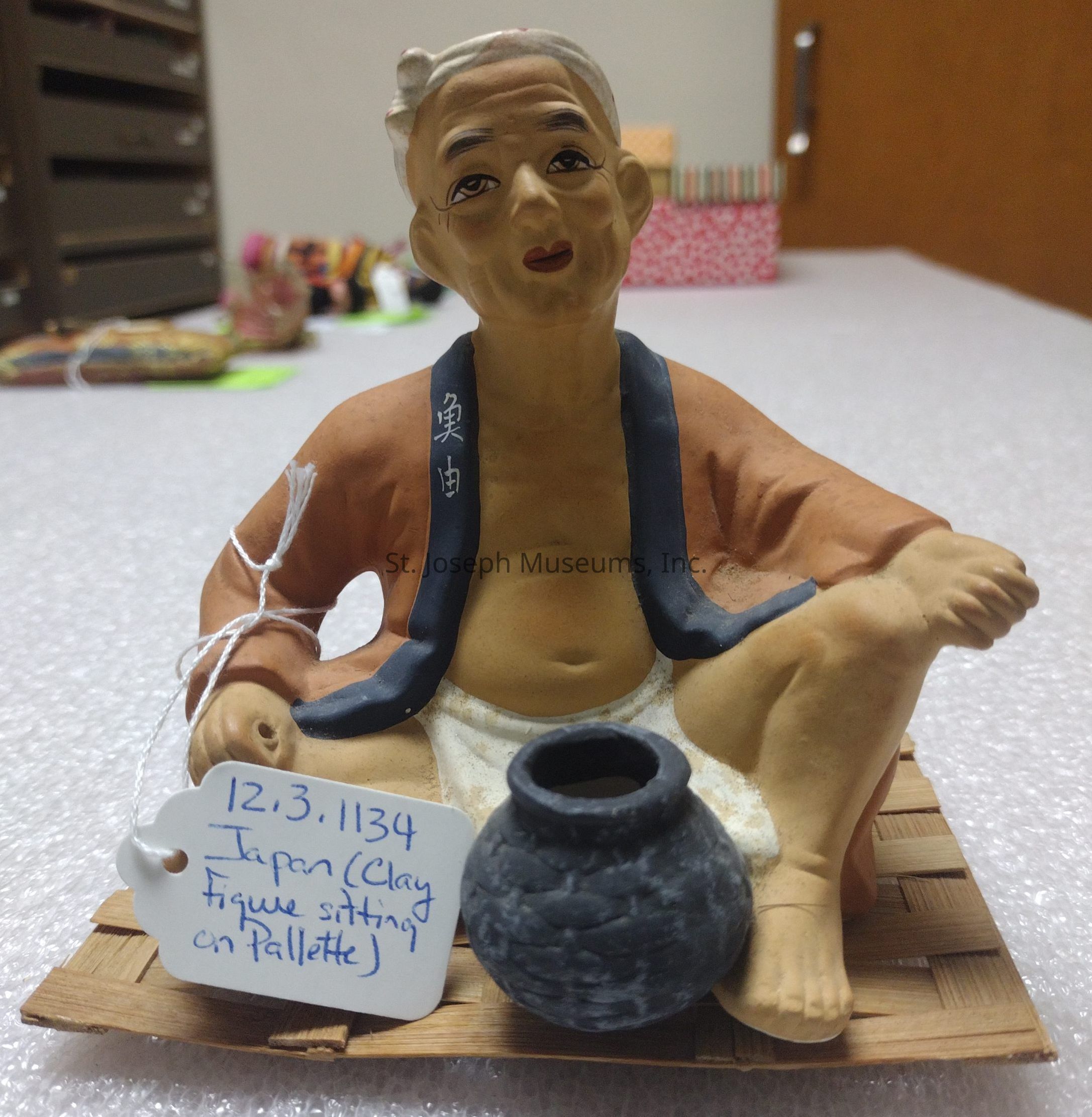The image depicts a clay figurine of a seated man, crafted with intricate details, sitting on a small wooden palette. The figure is garbed in traditional attire, with a dark blue collar and an orange robe, showcasing realistic facial expressions and body posture. Beside the figure, there is a textured gray pot, enhancing the artistic composition. Attached to the figurine is a tag labeled "12.3.1134 Japan (Clay figure sitting on pallet)," providing contextual information about its origin. The background reveals a room with various objects blurred in the distance, adding depth to the scene.