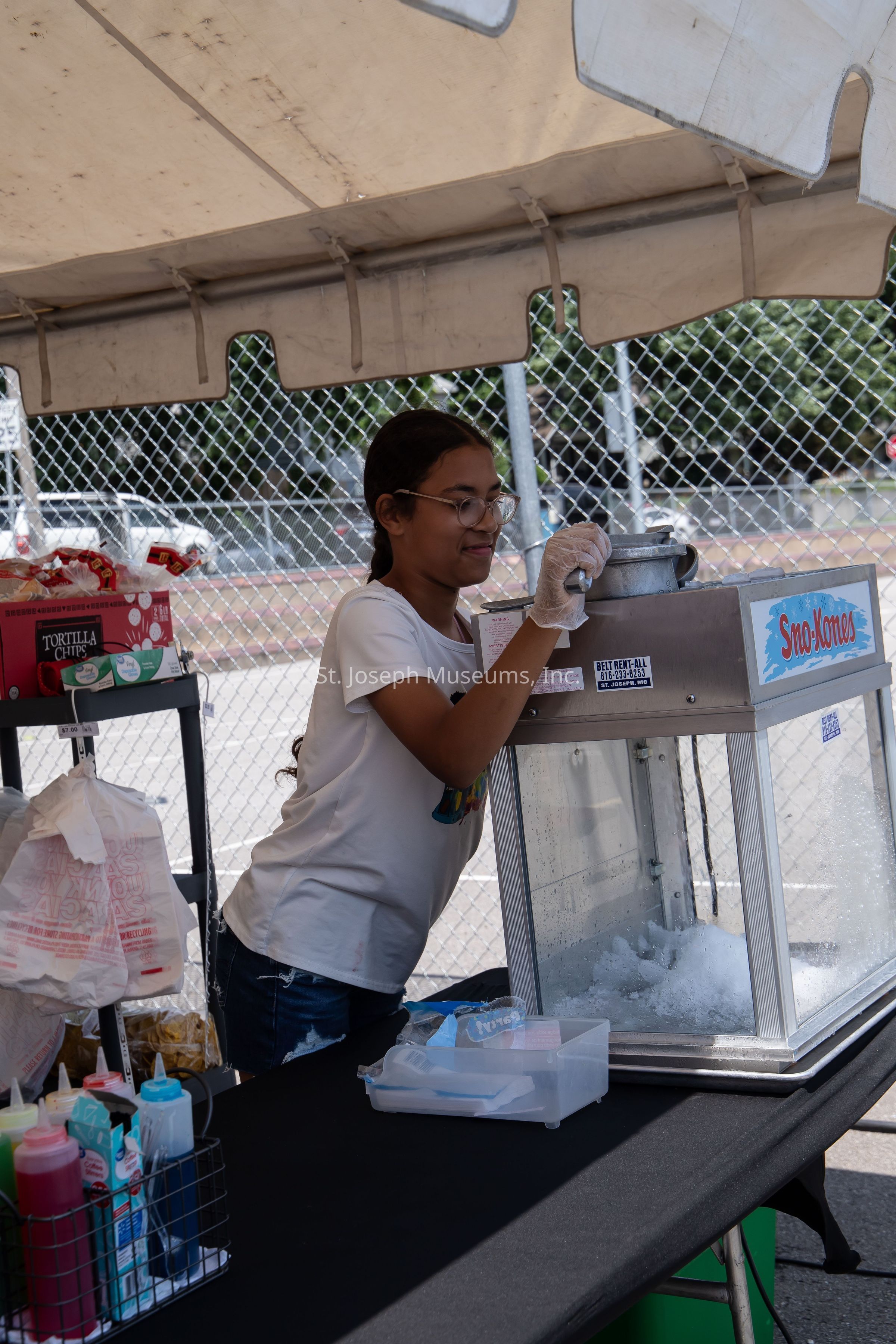 Beneath the shade of a canopy tent, a young girl is operating a snow cone machine at an outdoor event, using an ice grinder to prepare the frozen treat with focus and enjoyment. She is wearing a white t-shirt and glasses, with her hair tied back, and is protected by gloves for hygiene. The table in front of her is organized with various supplies and colorful syrup bottles ready for application on the snow cones. To her left, there are bags of tortilla chips and a collection of supplies neatly arranged in a box. Behind her, a chain-link fence separates the area from a parking lot or street, where a car is barely visible. The entire setup suggests a casual and friendly environment, possibly a community fair or local festival, where simple pleasures like snow cones bring delight on a warm day. The focus and enjoyment on the girl's face leave an impression of her diligence and the satisfaction of serving refreshing treats to attendees.
