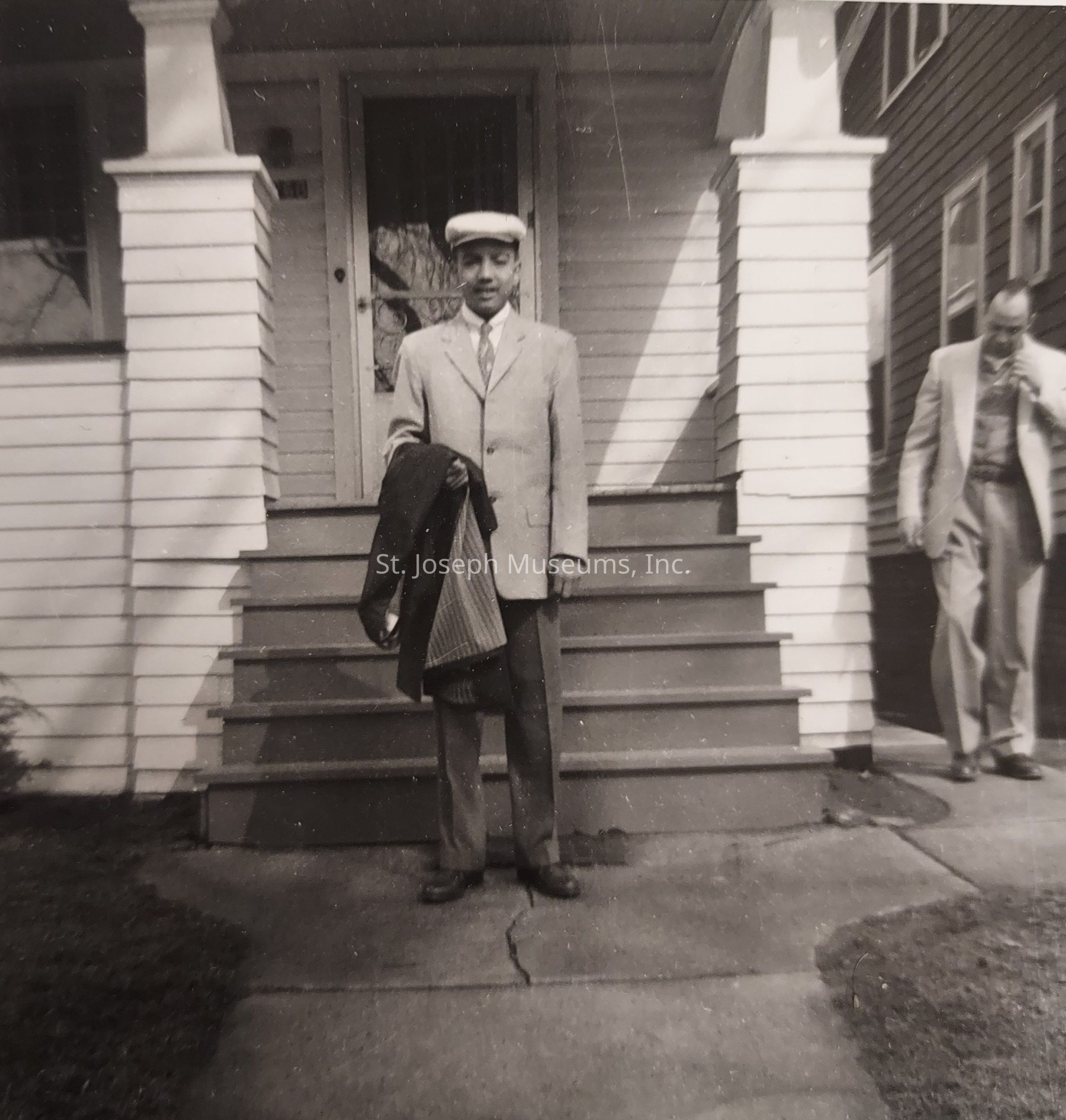 In the black and white photograph, a man stands confidently at the forefront, dressed in a neatly tailored suit, holding a coat in one hand while wearing a flat cap that complements his formal attire, and behind him is a house with distinct wooden siding and steps leading to the entrance, capturing the timeless style of mid-20th-century architecture; another person, also clad in a suit, appears to be descending the stairs, adding a sense of motion and balance to the scene, which is set against a backdrop that suggests a calm, possibly suburban environment.