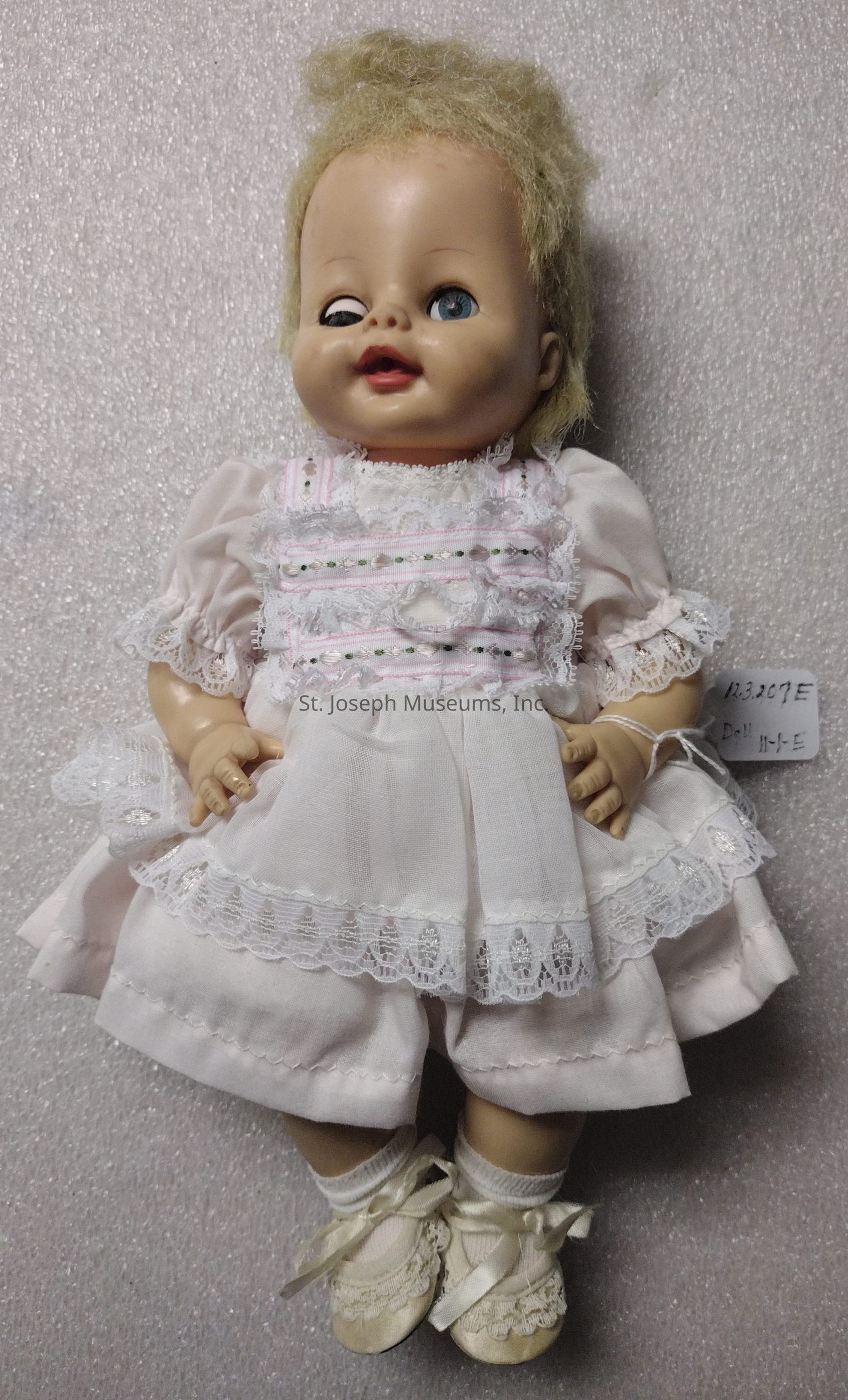 The image shows a vintage baby doll with a slightly weathered appearance, featuring short, light-colored hair that is somewhat disheveled. The doll has a pair of bright blue eyes, with one being slightly askew, giving it a whimsical expression. It is dressed in a delicate, lacy white dress with intricate detailing, including pink and green embroidery across the front, and matching white lace trim around the sleeves and hem. The outfit is completed with white bloomers underneath. On its feet, the doll wears small white socks paired with dainty white shoes, tied neatly with satin ribbons. A small tag can be seen attached to the doll’s wrist, inscribed with numbers and letters, though the purpose of the tag is not immediately clear. The background of the image is a textured, light-colored surface that provides a neutral backdrop, highlighting the doll's features and attire. The overall vibe of the scene is charmingly nostalgic, evoking memories of classic childhood toys.