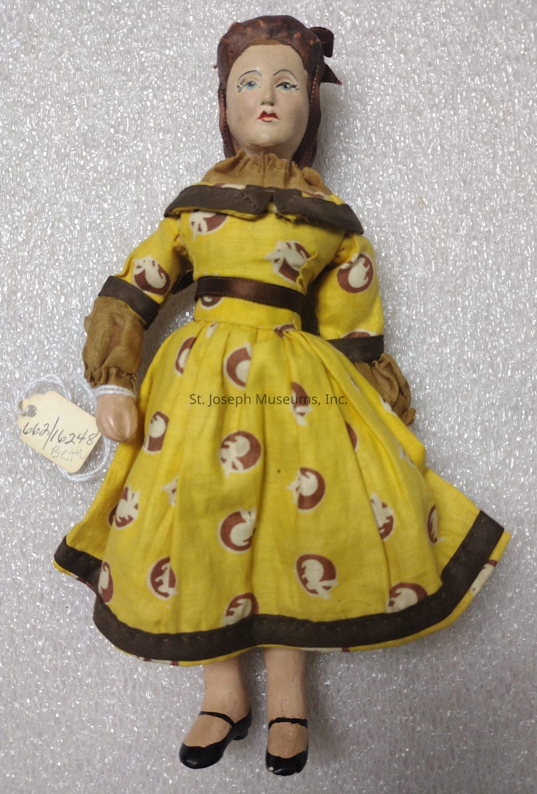 The image depicts a vintage doll dressed in a vibrant yellow dress featuring a pattern of circular brown and white designs. The dress is adorned with a dark brown trim on the collar, sleeves, and hem, complementing the doll's neatly painted black shoes. The doll's facial features are delicately hand-painted, with distinct blue eyes and red lips. Her hair is styled in an elegant updo, providing a classical appearance. Attached to her left hand is a small tag with handwritten text, indicating possible identification or cataloging information. The backdrop is a textured white surface, emphasizing the vivid colors of the doll’s attire and enhancing its nostalgic charm.