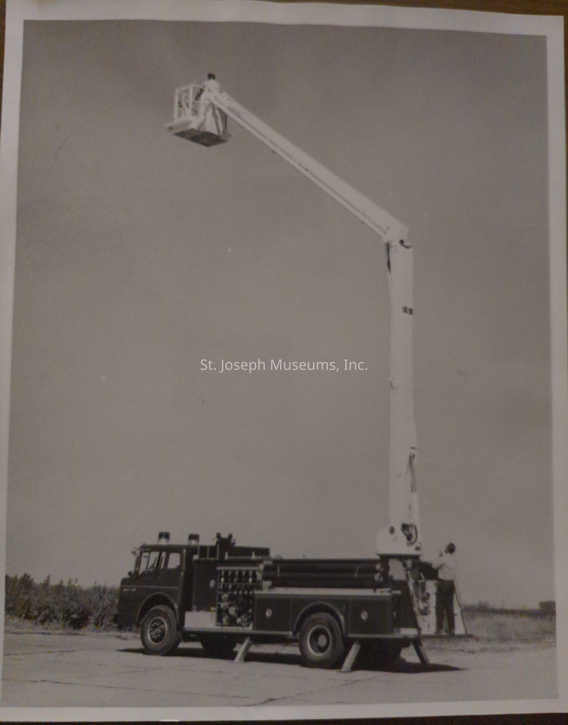 Black-and-white photograph depicting a fire truck from Snorkel Co., positioned on Lake Road and Arizona, featuring an extended aerial platform. A person operates the platform high above, showcasing its impressive reach and functionality, with the truck placed on a flat road, illustrating its readiness for firefighting operations.
