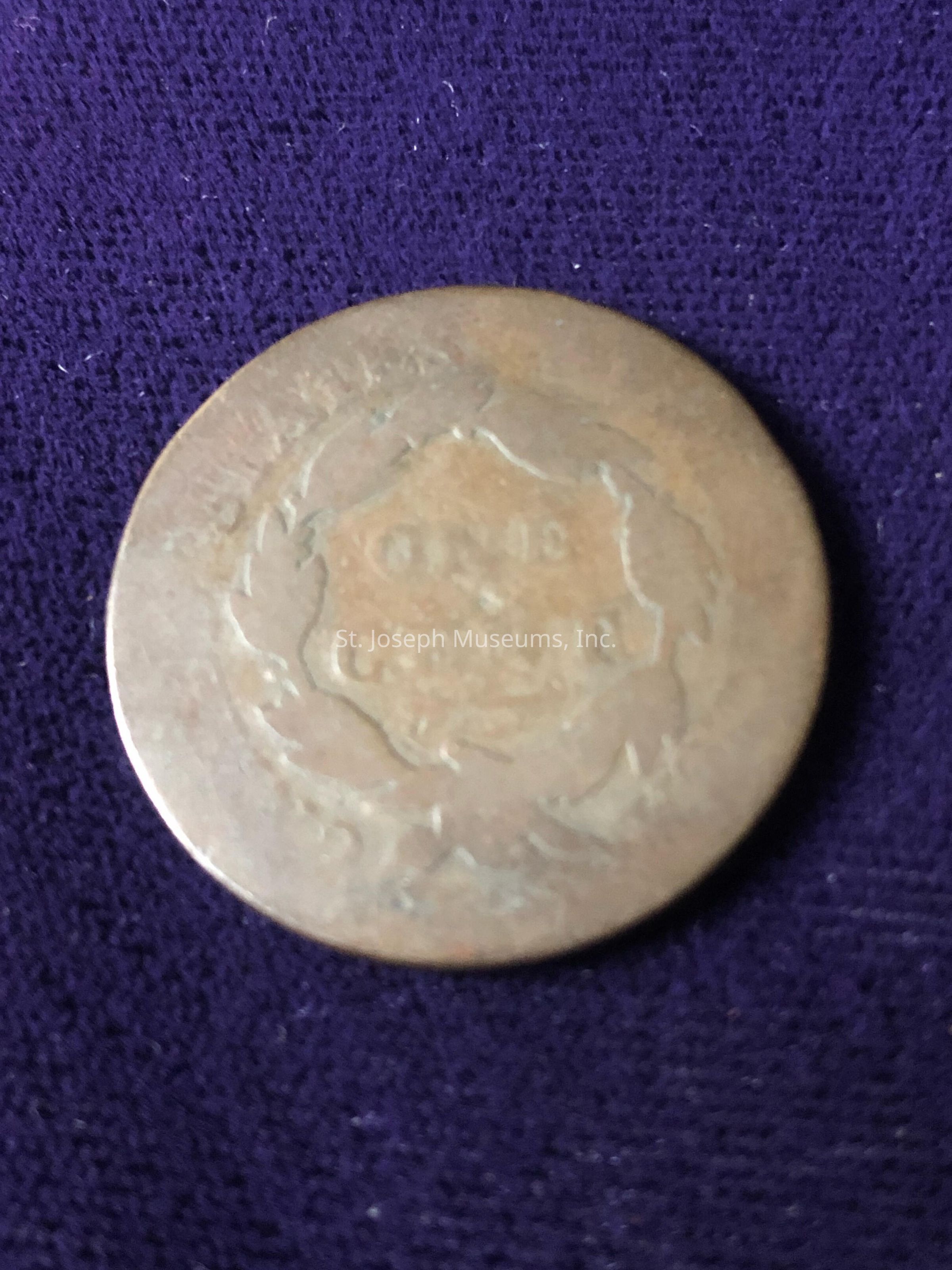 The coin surface exhibits noticeable wear and patina, characteristic of aged metal, with indistinct engravings that are challenging to discern due to its condition; these markings appear to include some form of text or emblem, possibly enclosed by what might be a wreath or laurel design; the blurred and slightly reflective surface hints at its prolonged exposure or contact over time, suggesting this coin could hold historical or numismatic value to someone familiar with its origin and context.
