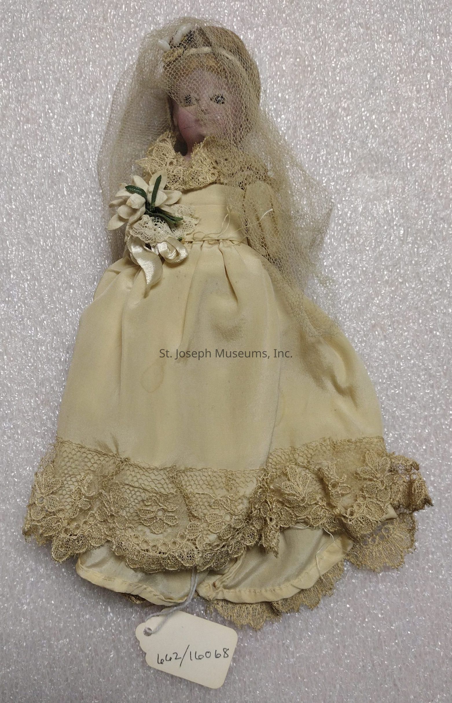 The image depicts a vintage doll dressed in a wedding ensemble. The doll is adorned in a cream-colored gown with intricate lace detailing at the hem and collar. A delicate veil, crafted from fine netting, covers the doll's head, adding to the bridal theme. The bodice is accentuated with small fabric flowers and ribbons. The doll's facial features include rosy cheeks and painted eyes, and its hair is styled neatly underneath the veil. A tag attached to the doll bears the handwritten numbers "662/16068," suggesting an identification or cataloging system. The textured background suggests a soft, possibly fabric surface, contributing to the overall antique and delicate aesthetic of the photograph.
