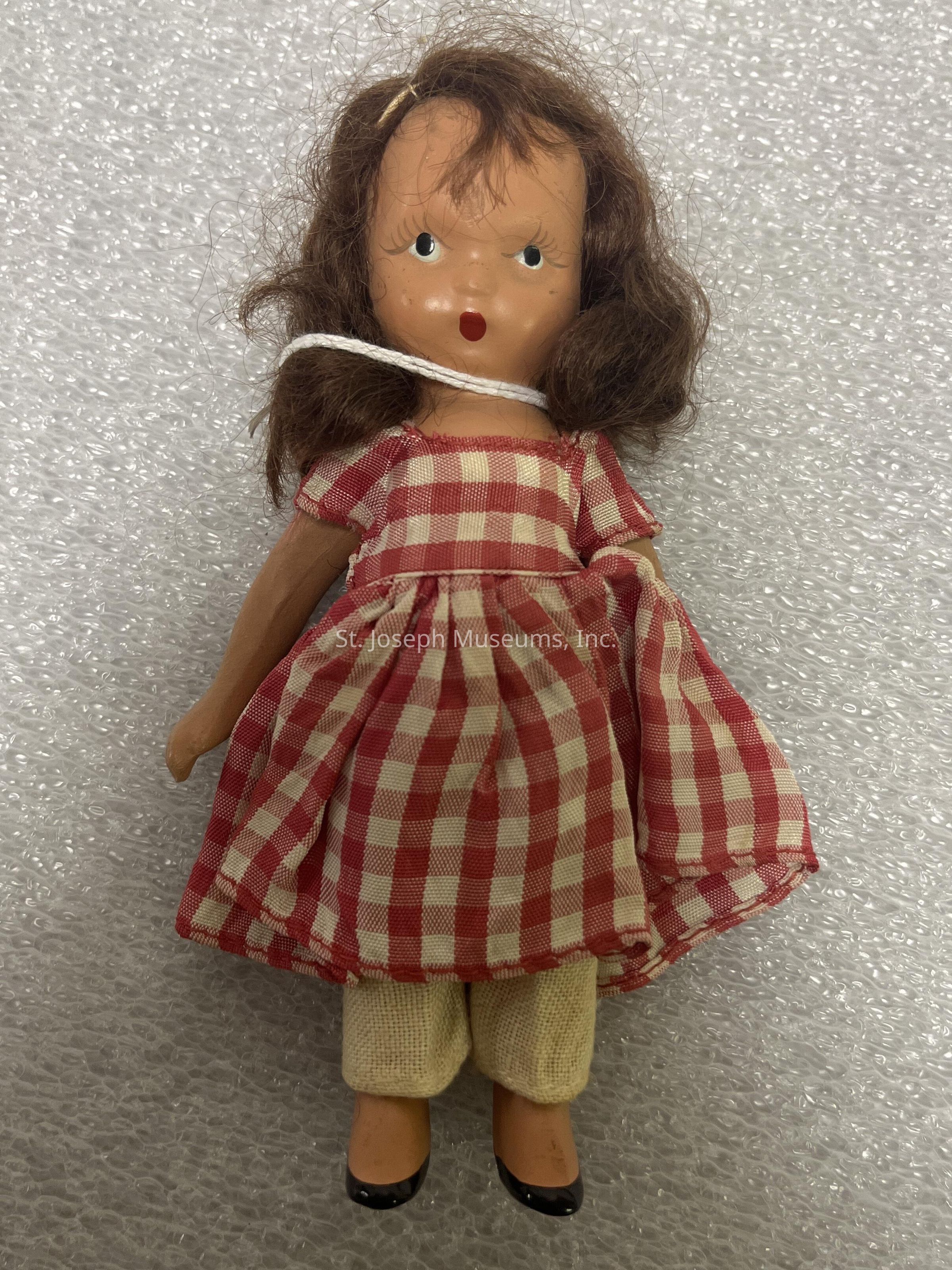 The image depicts a vintage doll with a whimsical expression, characterized by wide blue eyes and a small open mouth. The doll has curly brown hair and is wearing a red and white checkered dress which adds a charming, rustic appeal. Below the dress, she is dressed in cream-colored pants. The doll is standing against a textured, light background, possibly bubble wrap, which provides a contrasting backdrop to her attire. Her shoes are painted on, completing the quaint and nostalgic look that is reminiscent of mid-20th-century style.