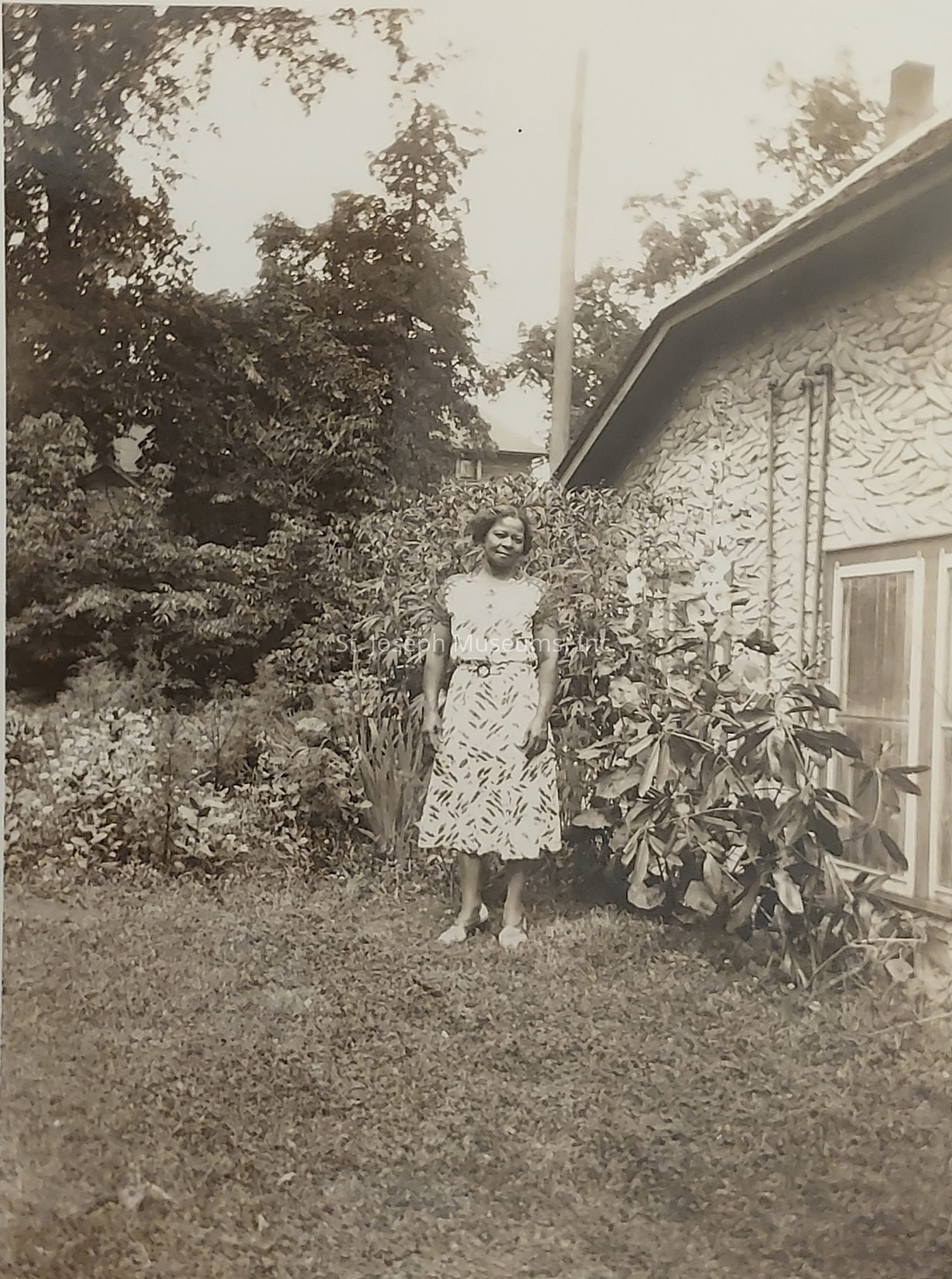 In this vintage black-and-white photograph, a woman stands in a garden beside a house with a textured exterior wall. She is wearing a patterned dress and smiling, conveying a serene presence. The garden is lush with various plants and bushes, creating a picturesque backdrop. Tall trees extend towards the sky in the background, enhancing the natural setting. The house has a prominent window, suggesting a cozy and welcoming home environment. This image captures a moment of tranquility and simplicity, evoking a nostalgic sense of peace and timelessness.