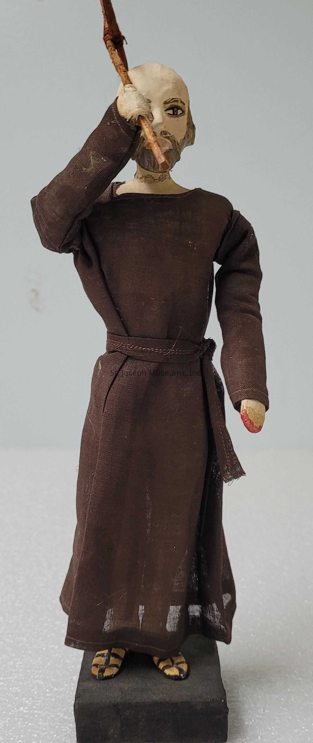 The image depicts a handcrafted wooden figure dressed in a dark brown robe tied with a simple belt at the waist. The figure appears to be in a contemplative pose, raising one hand to its face, while holding a stick in the other hand. Its facial features are rudimentary but expressive, suggesting a thoughtful or introspective mood. The figure stands on a black rectangular base, and its feet are covered with striped sandals. The craftsmanship exhibits a rustic charm, with visible textures in the fabric and wood, providing the figure with a humble and monastic appearance.