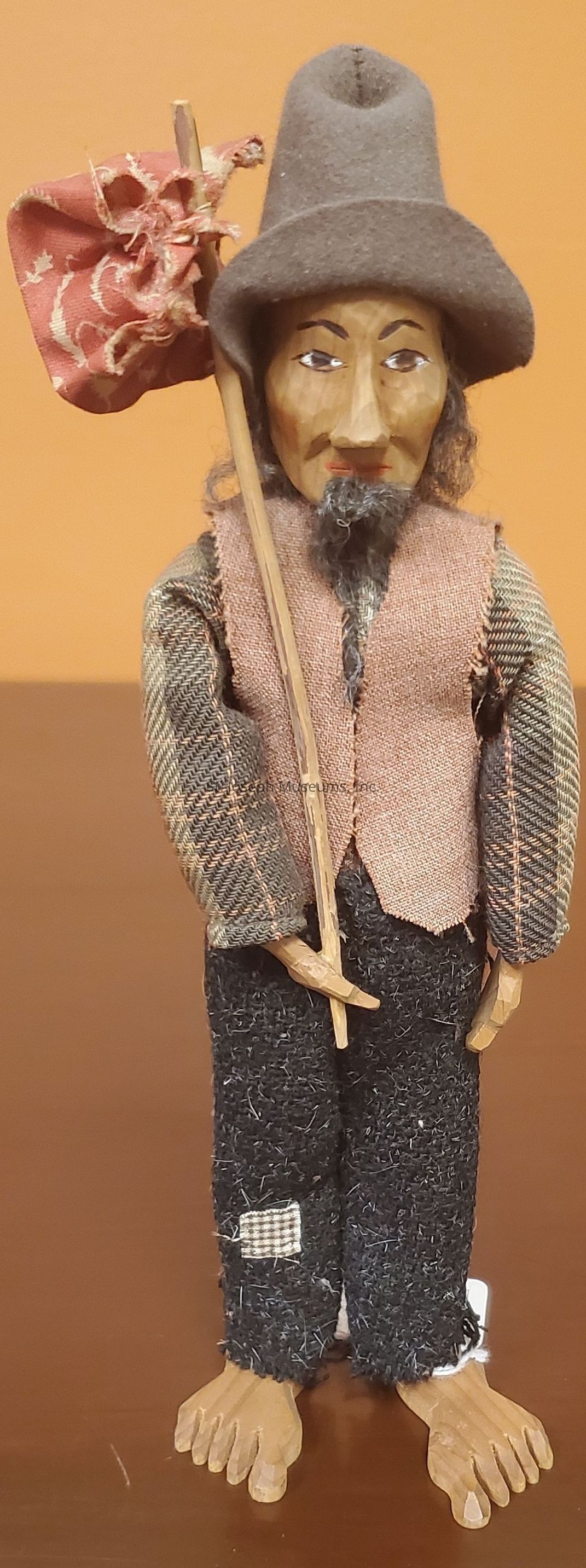 The image showcases a handmade wooden figurine depicting a man with a detailed, expressive carved face, complete with a modest beard and mustache, giving him an aged appearance. He is dressed in a rustic, artisanal outfit consisting of a brown textured vest layered over patterned long-sleeved attire, indicating a rural or traditional setting. His trousers are dark with a patch on one leg, adding to the handcrafted, vintage aesthetic. The figurine is barefoot, and on his head rests a large, soft fabric hat reminiscent of old rural headwear. He holds a stick in his right hand, where a small, worn red flag with subtle decorative patterns is attached, flowing at one corner. The background reveals a plain, warm-toned setting, enhancing the earthy, traditional vibe of the entire piece, emphasizing the craftsmanship evident in its creation.