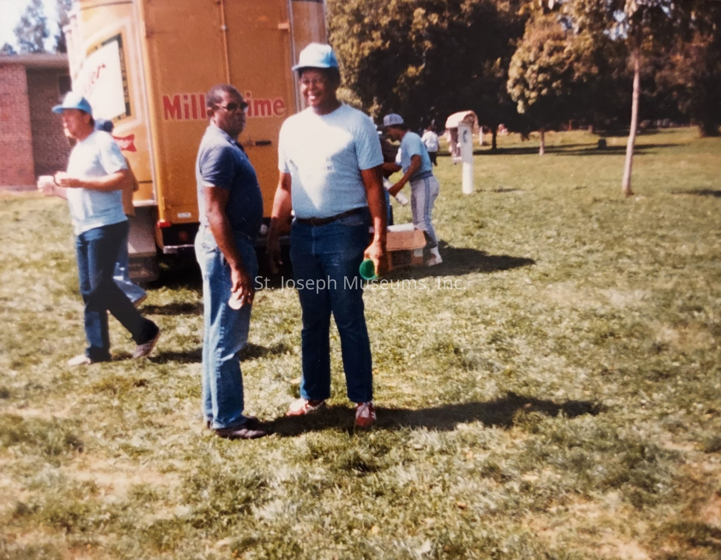 In the image, there are four individuals outdoors on a grassy area. Two of them, standing front and center, are smiling and appear to be engaged in conversation. One holds a green cup, while the other has their hands by their side. Both are casually dressed in jeans and light blue shirts, with one wearing a cap. In the background, near a yellow truck with red text, another person holds a cup, wearing a similar outfit. Further behind, a fourth person interacts with a box on the ground, as trees and an open area expand around them, indicating a park setting on a sunny day.