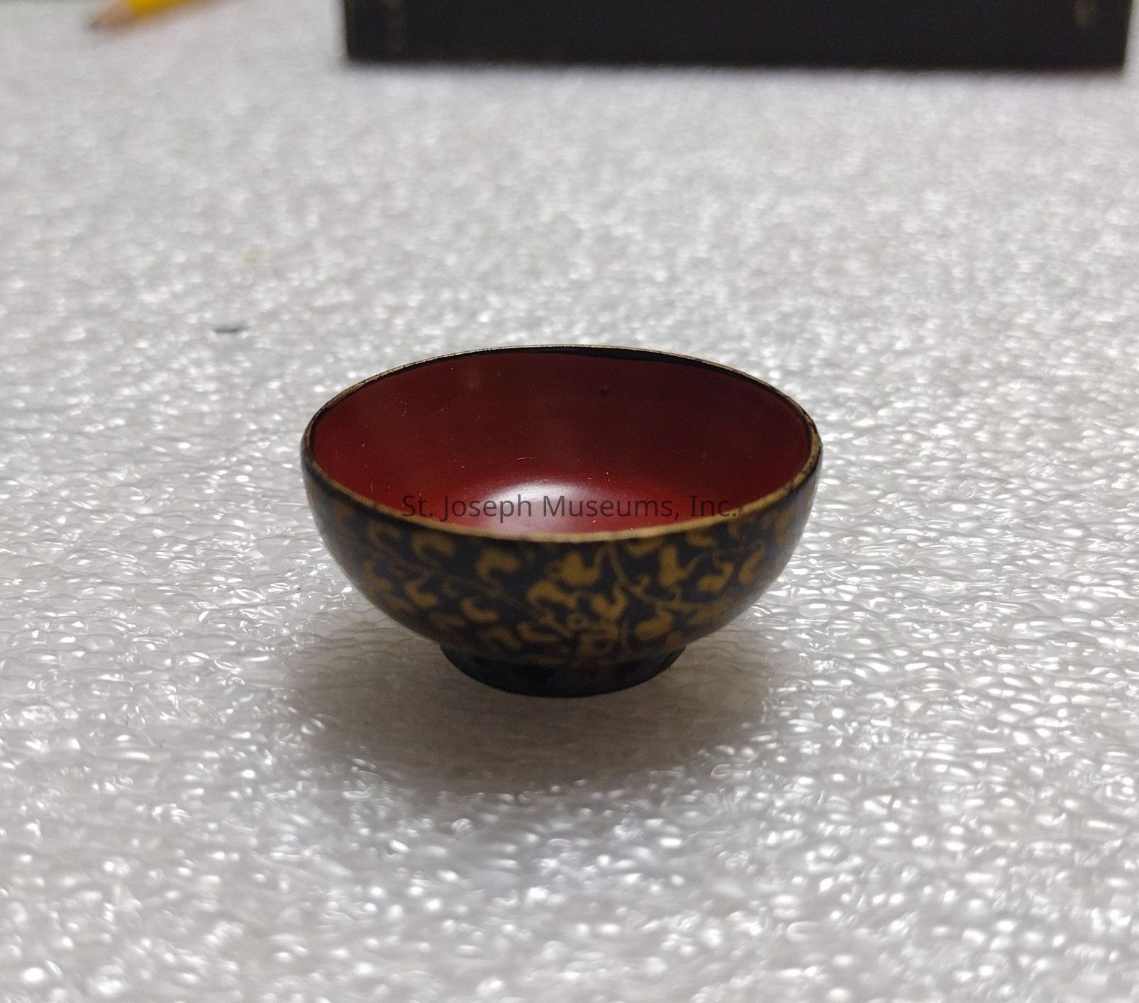 The image depicts a small, intricately decorated bowl resting on a surface covered with a bubble wrap. The bowl features a deep red interior, which appears smooth and glossy, while its exterior is adorned with an ornate pattern in gold, creating a striking contrast against its dark background. Its circular shape and shallow depth suggest it may be used for special purposes, possibly holding small quantities of liquid or decorative items. The light source from above casts gentle reflections on its surface, enhancing the elegance and craftsmanship of the bowl. In the background, a subtle blur is present, drawing focus to the craftsmanship of the bowl itself, while additional elements like the blurred pencil add context to the setting, hinting at a casual or working environment where such an item might be admired or utilized.