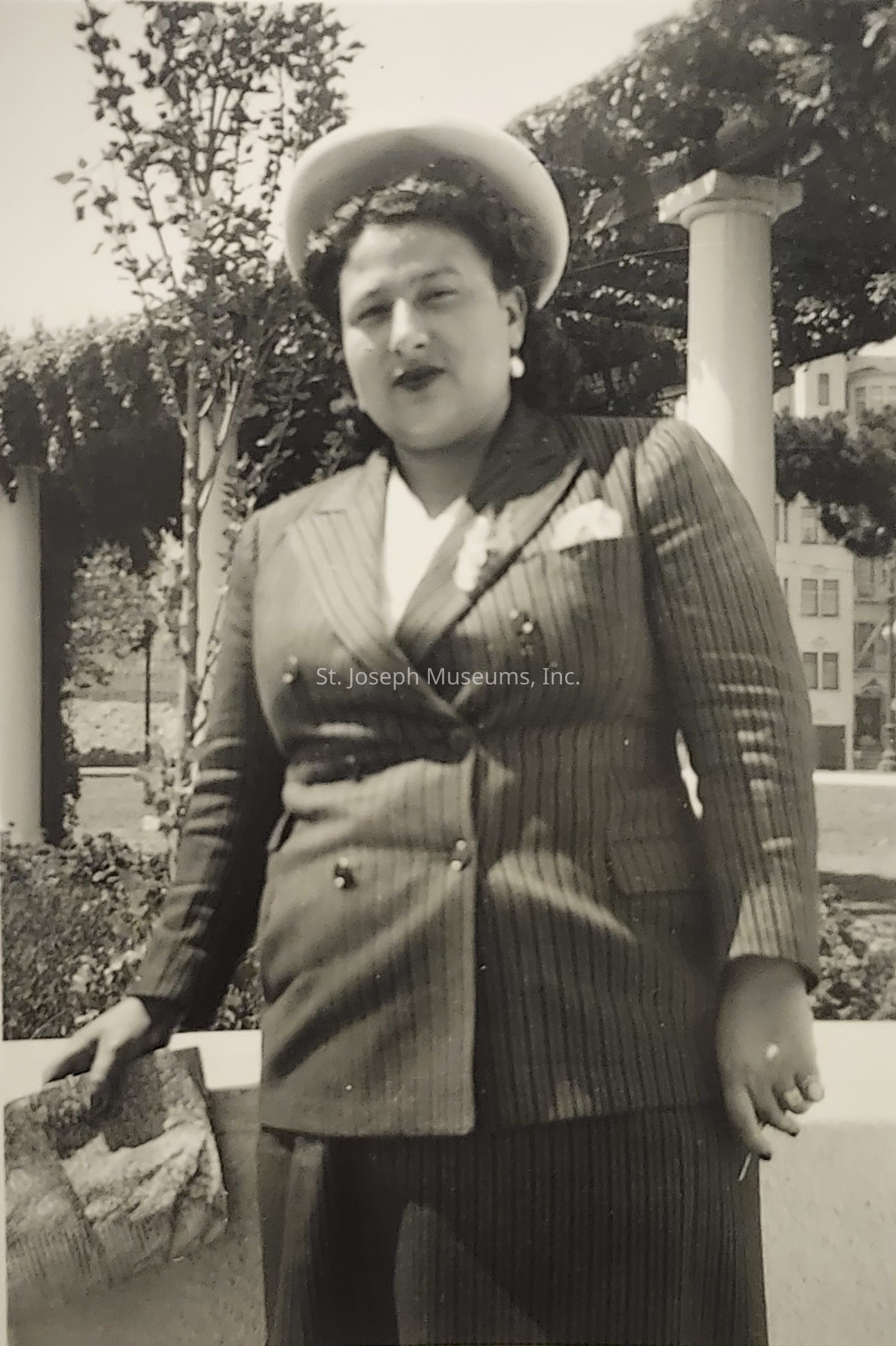 An elegantly dressed woman stands confidently in a vintage black-and-white photograph, wearing a pinstriped, double-breasted suit with a white shirt underneath, accessorized with a stylish hat perched on her wavy hair and a small, textured clutch in her left hand. The background features a beautifully landscaped garden area with neatly trimmed foliage and towering trees, framed by white columns that add a classic touch to the scene. The woman's expression is calm and composed, adding dignity to her poised stance.