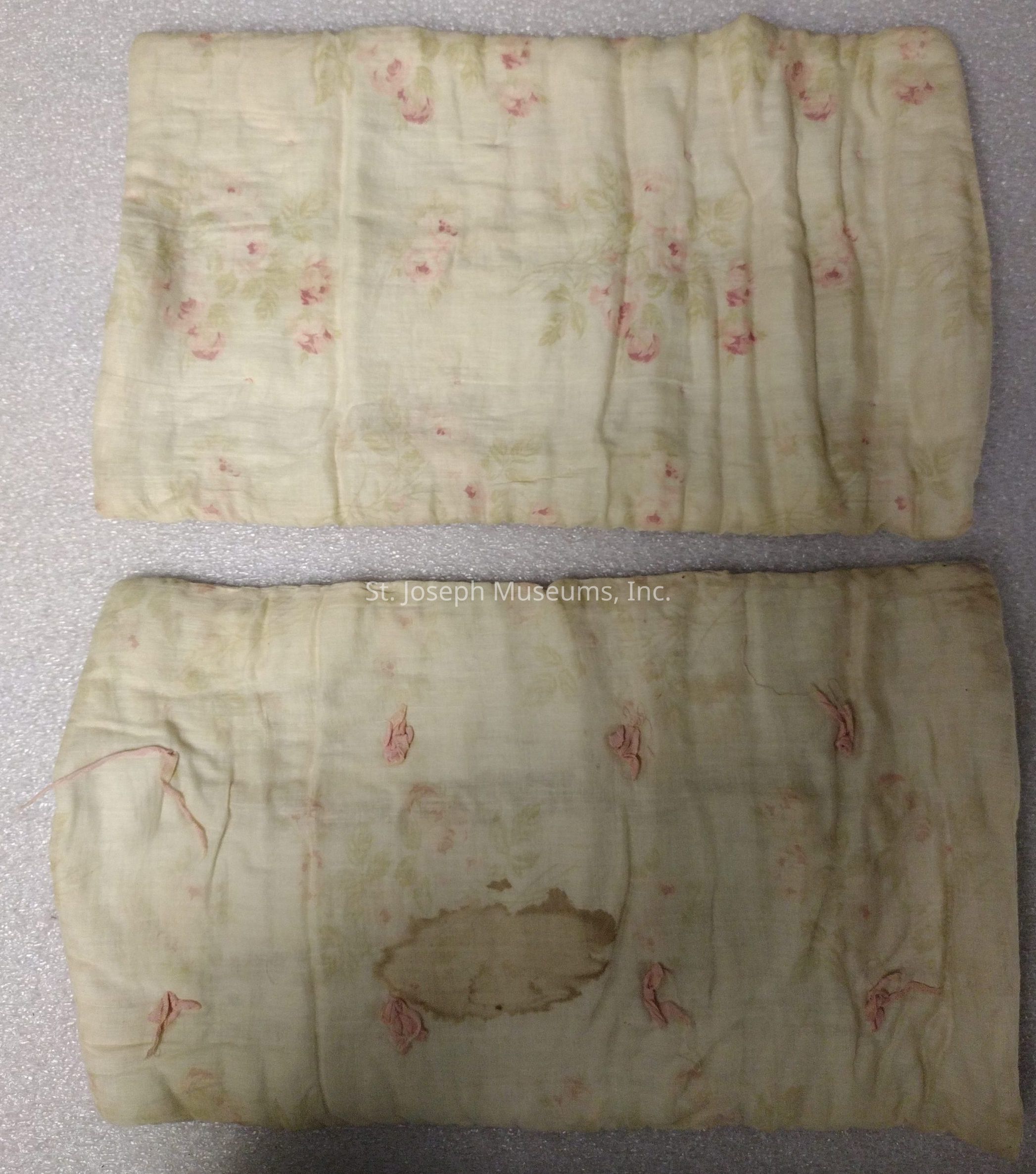 This image shows two pieces of vintage fabric laid out on a textured, light-colored surface. The fabric appears to be aged and has a light cream background adorned with a delicate floral pattern containing small pink flowers and subtle green leaves. The top piece is relatively smooth with gentle folds, while the bottom piece exhibits noticeable signs of aging, including a prominent brown stain toward the center and some unraveling at the edges. Both pieces share a similar color palette and design, suggesting they are part of the same set or material.