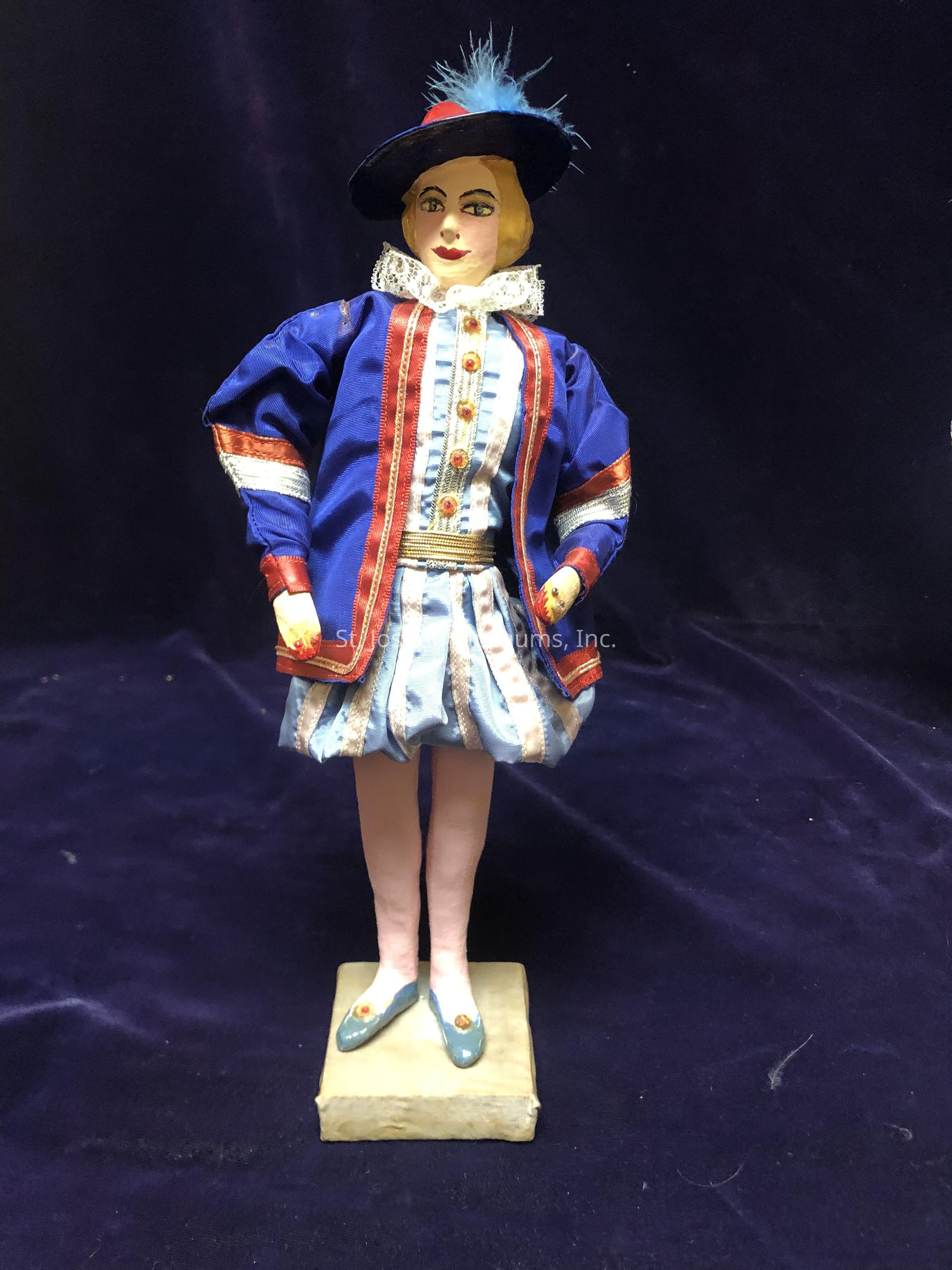 This image shows a detailed and colorful doll dressed in a historical outfit, featuring a vibrant blue ensemble with red and white accents. The doll is adorned with a ruffled collar and a large, feathered hat that matches the blue of the outfit. Its outfit includes a jacket and a skirt with intricate embellishments and gold trim, highlighting an era reminiscent of Renaissance or Elizabethan fashion. The doll stands on a small pedestal, and its shoes are painted in a complementary blue shade with decorative elements. The background is a dark, rich fabric, which enhances the vividness of the doll's attire and creates an elegant contrast, emphasizing the detailed craftsmanship of the doll's costume. The expression of the doll is serene, with painted features that suggest a delicate, poised presence, capturing a sense of historical elegance.