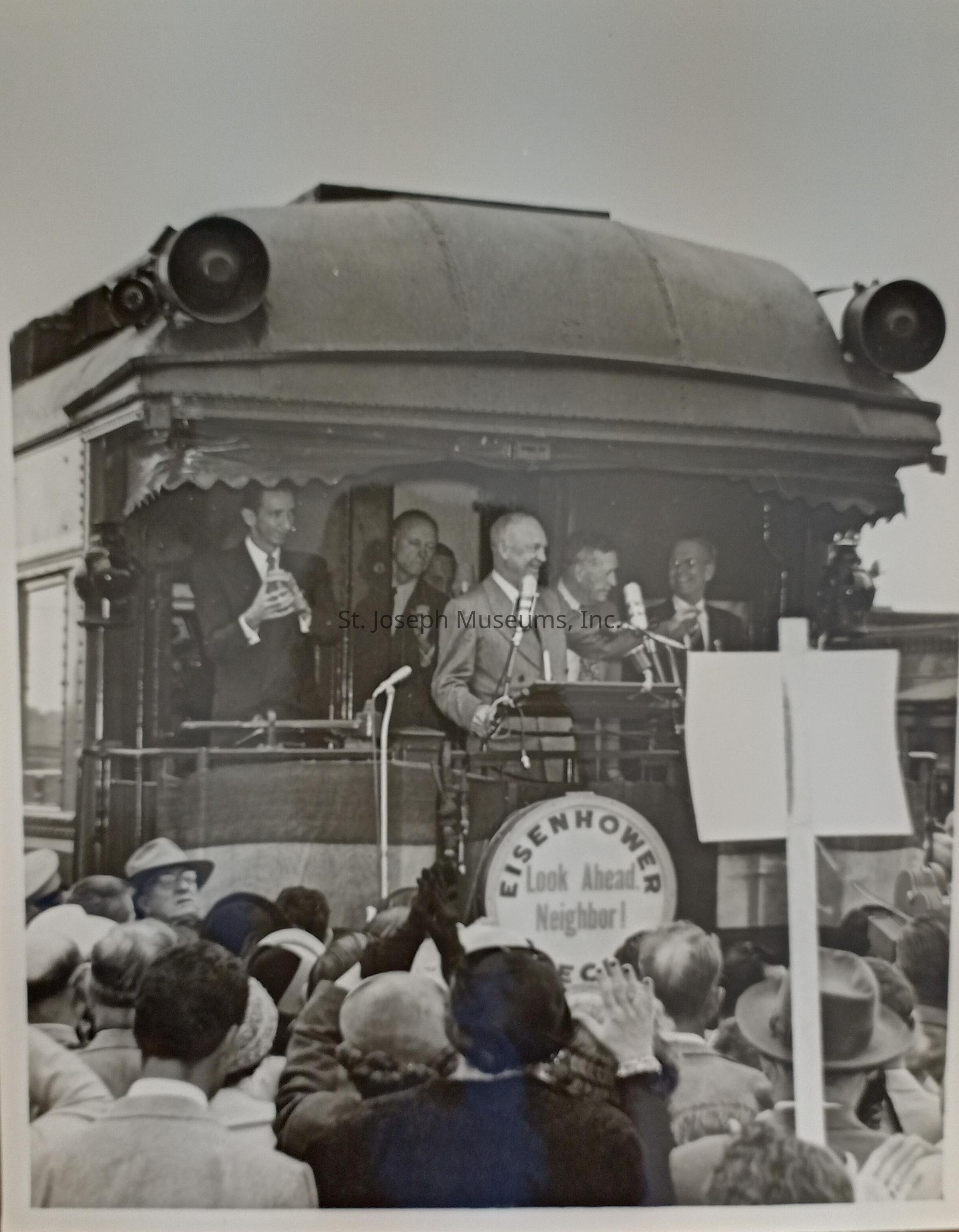 Dwight Eisenhower stands beside his wife, Mamie, on the platform of a caboose during a campaign stop in St. Joseph, both smiling warmly at the enthusiastic crowd. The onlookers eagerly reach out towards the couple, showing their support amid the "Eisenhower" banner that prominently displays the campaign slogan, "Look Ahead, Neighbor!"