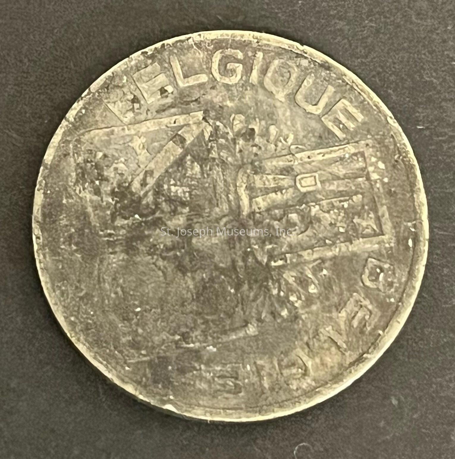 The image shows a worn coin displaying signs of age, with the text "BELGIQUE" visible at the top, indicating it is from Belgium. The surface is eroded, making details difficult to distinguish, but parts of the design, possibly a coat of arms or emblem, can still be seen. The coin's metallic surface appears tarnished, giving it a dull, weathered look. Its circular shape and remaining inscriptions suggest it's a currency piece, possibly historical. The background is a plain, dark surface, which contrasts with the lighter, more textured appearance of the coin. The coin's edge is slightly rough, adding to its vintage aesthetic.