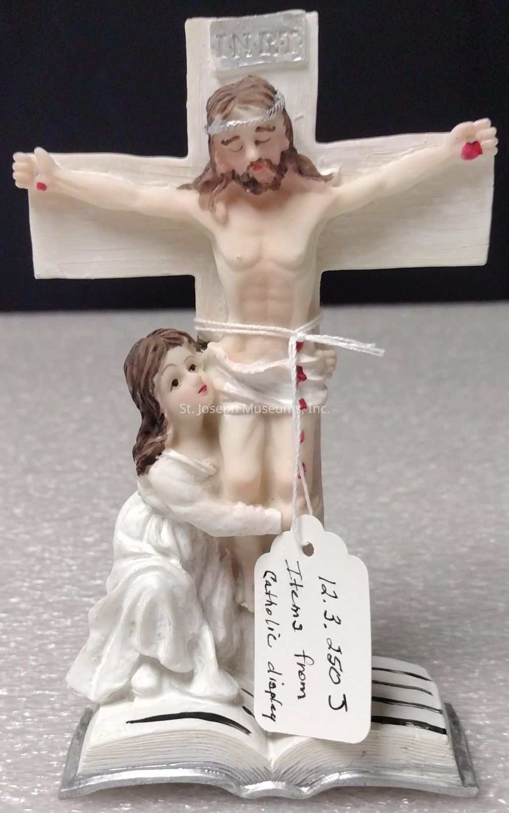 The image depicts a detailed religious figurine, featuring a depiction of Jesus on the cross, with the label "INRI" above his head. Below Jesus, a woman in a white robe is kneeling, embracing the base of the cross, with an expression of reverence. The figurine is set on an open book, and a string is tied around the cross with a handwritten tag that reads, "Items from Catholic display," along with some numerical notations. The intricate sculpting and painting reflect a sense of devotion, capturing an intimate and solemn moment within a religious context. The background is blurred, highlighting the central figures and lending focus to the ornate design and emotional elements of the piece.