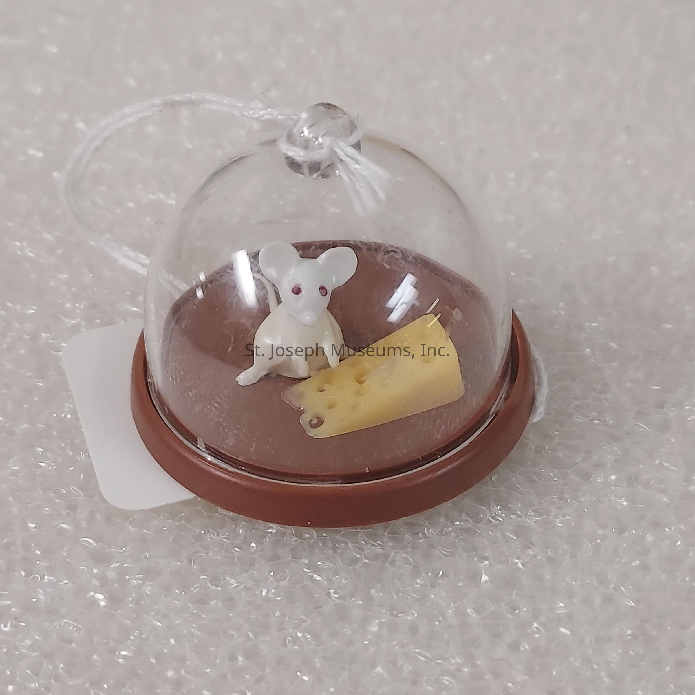 The image displays a small, decorative figurine consisting of a white mouse and a wedge of Swiss cheese, both encased under a transparent glass dome. The dome is situated on a brown circular base, and there is a white string attached to the top of the dome, possibly for hanging or display purposes. This charming miniature scene, with the mouse positioned attentively next to the cheese, is set against a textured, bubble-wrap-like background, which enhances its delicate appearance. The whimsical design suggests that it is a collectible ornament or a novelty keepsake.